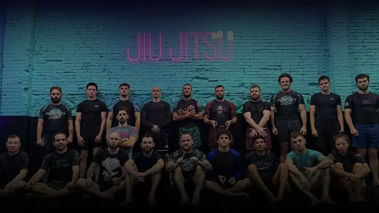 Group of people, including men and women, posing for a photo in front of a wall with a neon sign that reads 'JIU JITSU'