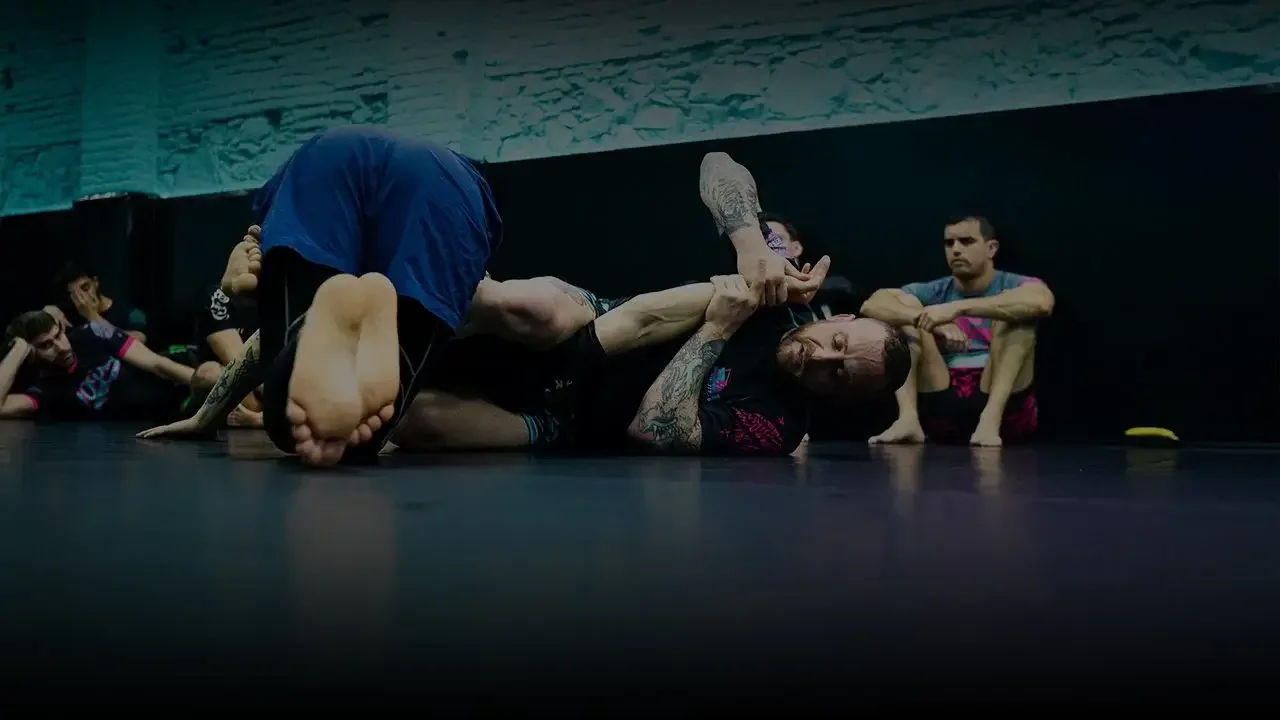 Two men practicing Brazilian Jiu-Jitsu on a black mat, with one applying a submission hold while the other tries to defend, and others observing in the background.