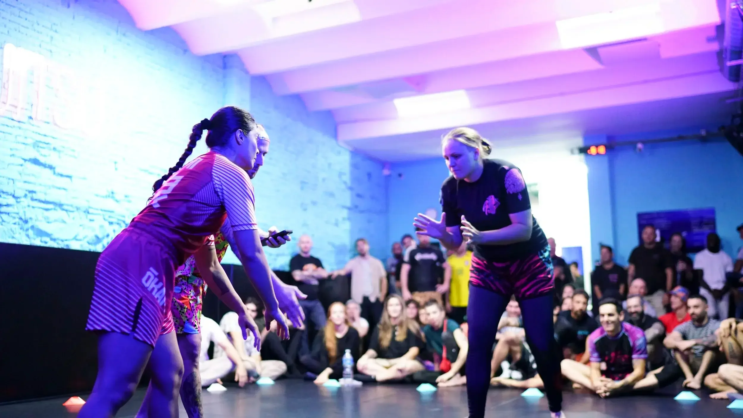 Two women engaged in a martial arts demonstration or sparring match in front of a seated crowd at an indoor event, with colorful lighting and a brick wall in the background.