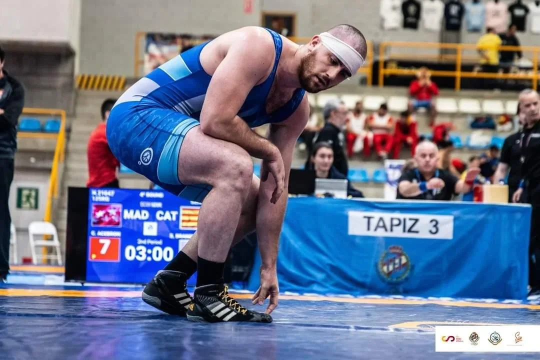 A male wrestler in a blue singlet and white headband crouches on a wrestling mat in a sports arena, with spectators and officials watching.