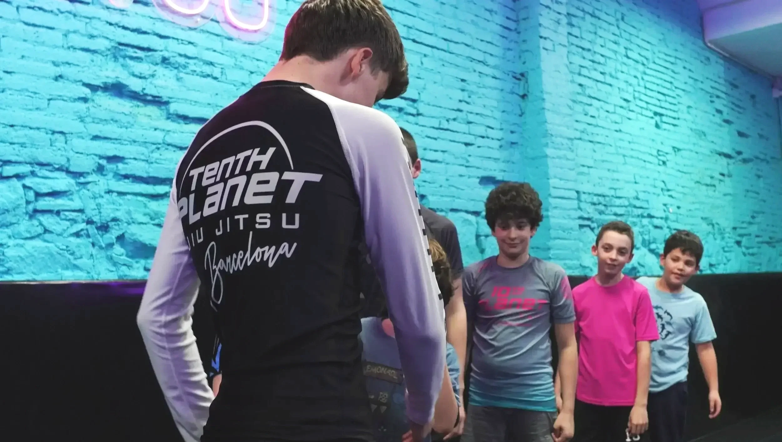 A group of children and a teenager with curly hair in a room with a blue brick wall and purple neon lights. The teenager is wearing a black and white sports jersey with "Tenth Planet Jiu Jitsu Barcelona" written on it. Some children are wearing colorful t-shirts, and they are standing and looking at the teenager.