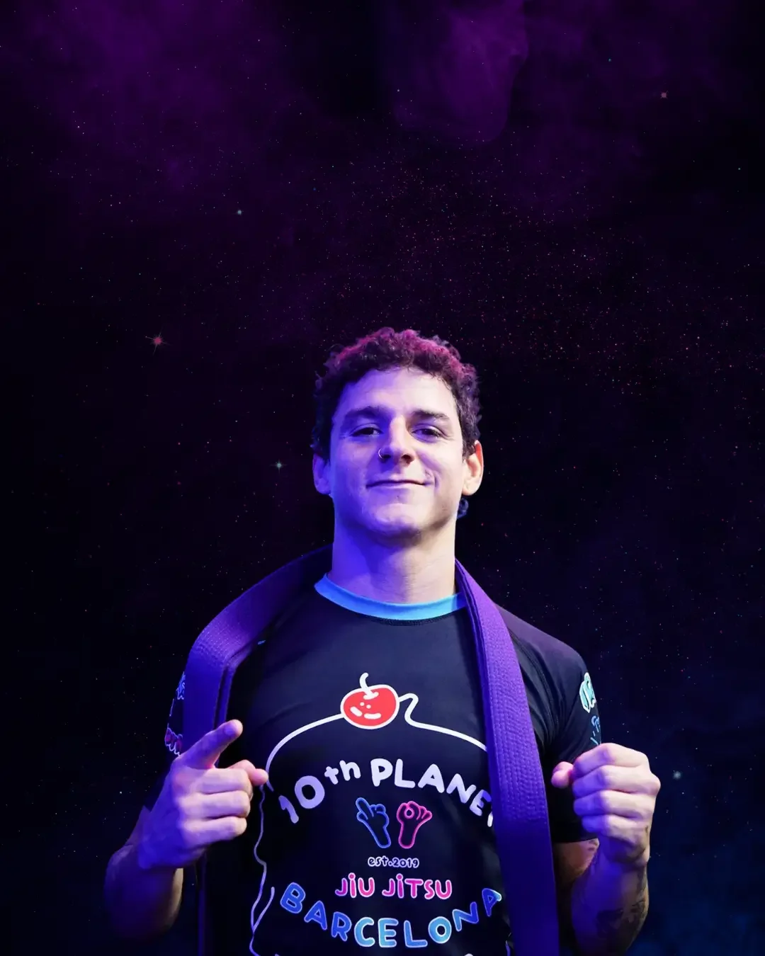 A young man in a black T-shirt with colorful text and graphics, smiling and holding a purple backpack, standing in front of a cosmic background with stars and purple nebulae.