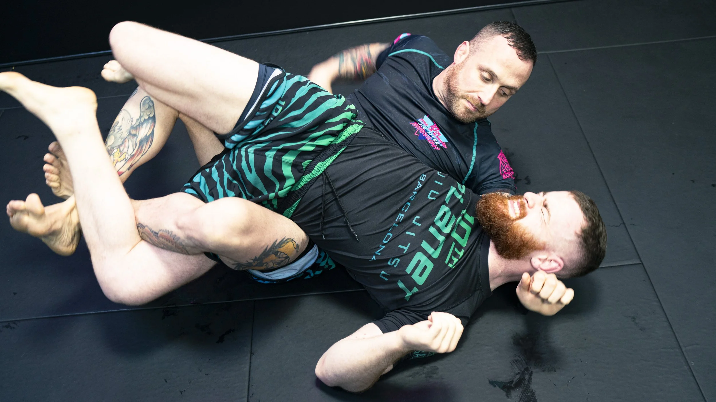 Two men are practicing Brazilian Jiu-Jitsu on a black mat, with one man in a dominant position and the other trying to defend.