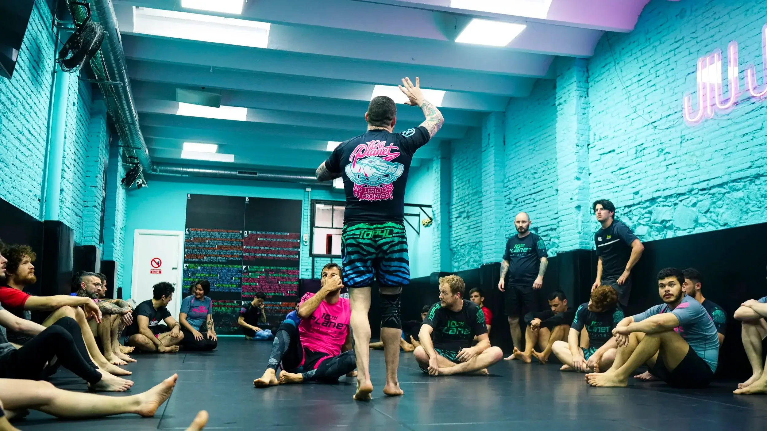 A group of people sitting on the floor inside a gym with blue brick walls and a neon sign, listening to an instructor who is demonstrating a move. The instructor is standing with his back to the camera, wearing a black shirt and colorful shorts. Some participants are sitting cross-legged, watching attentively.