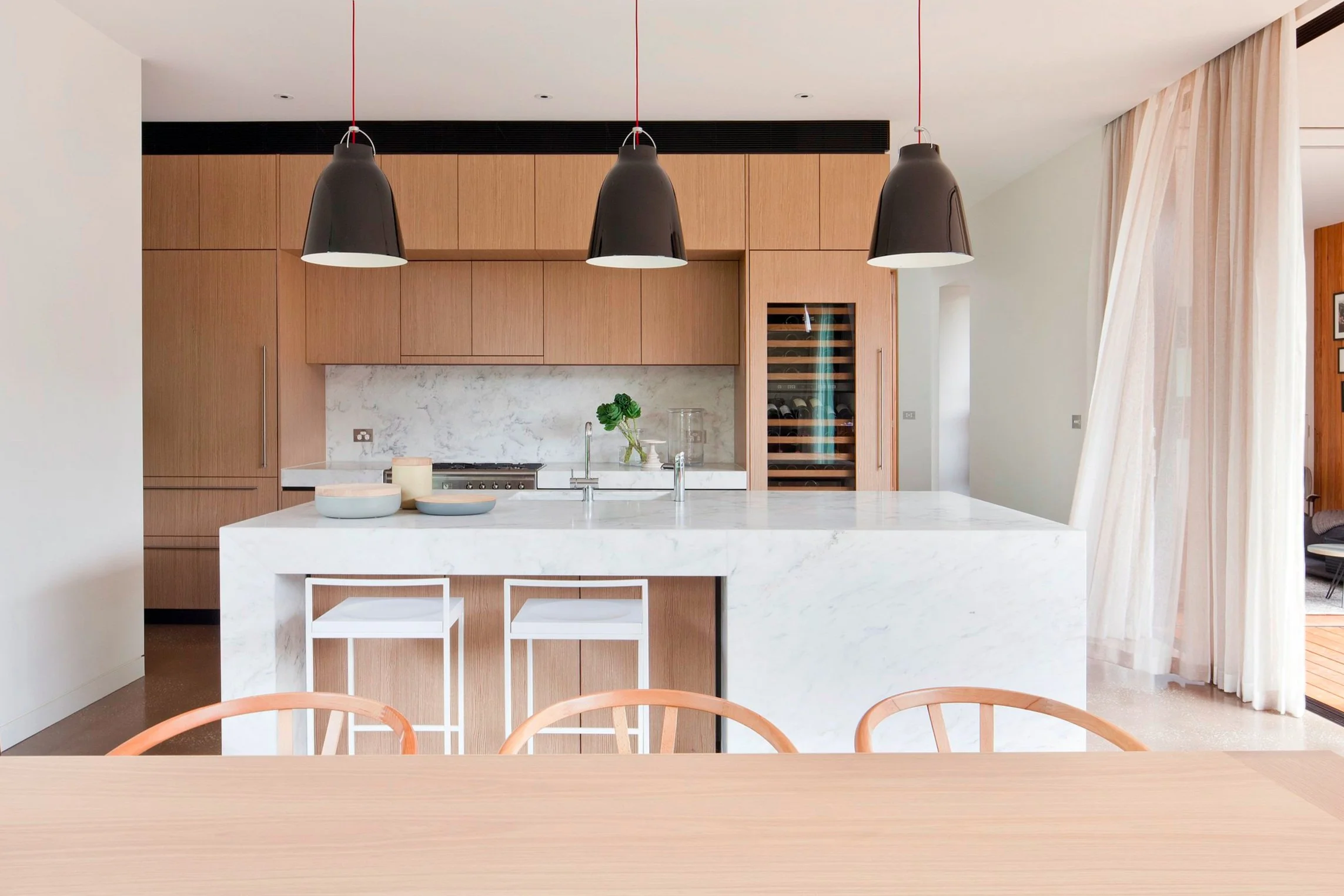 Moderne Küche mit einer Kücheninsel aus weißem Calacatta-Marmor, schwarzen Pendelleuchten, holzfurnierten Küchenschränken, einem eingebauten Weinschrank und Vorhängen vor einer großen Fensterfront.