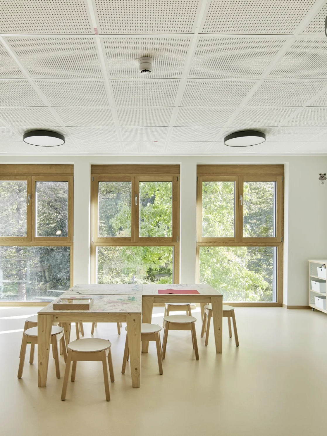 Schöner, heller Raum mit beigem Linoleumboden und großen, bodentiefen Fenstern, davor Holztische und Stühle für Kindergartenkinder.