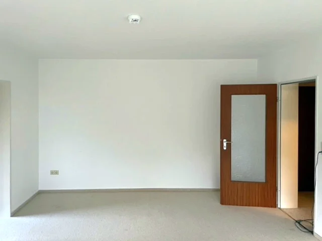 Eine leeres Wohnzimmer vor der Umgestaltung mit weißen Wänden und grauem Boden und einer affenstehenden Tür aus Holz.