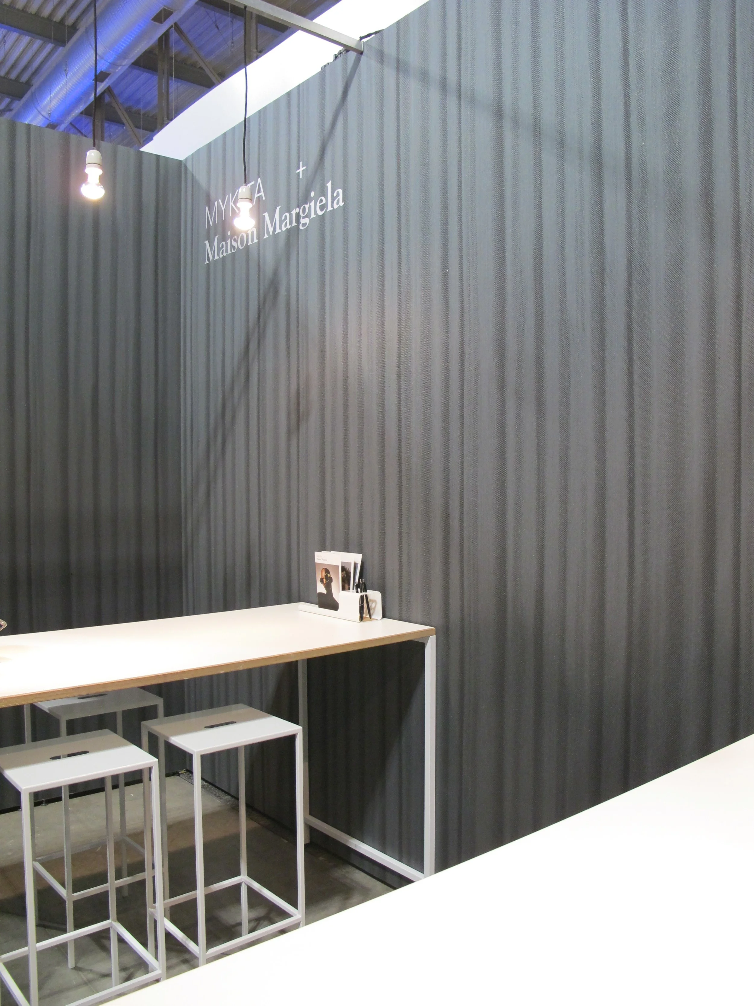 Exklusiver Verkaufsbereich auf dem Messestand mit weißen Tischen und Hockern, die die CI-Farbe des Unternehmens widerspiegeln. Die Wände sind aufwendig mit hochwertiger Tapete in Vorhangoptik verkleidet.