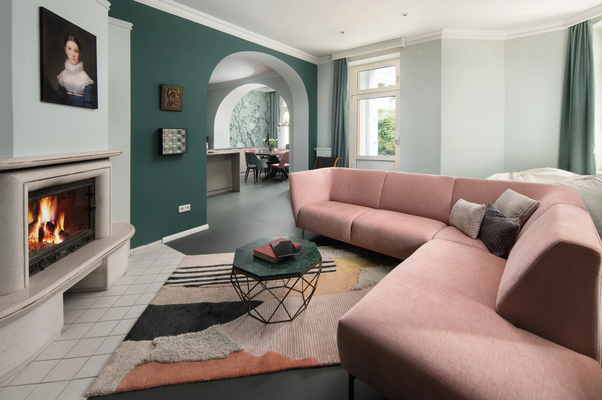 Gemütliches Wohnzimmer im Wohnstil Modern Glamour mit einem großen, rosafarbenen Sofa vor einem Kamin, einem bunten Teppich, großen Fenstern mit grünen Samtvorhängen, offener Blick in den Essbereich mit Esstisch und Stühlen. Die Wände in Minztönen.