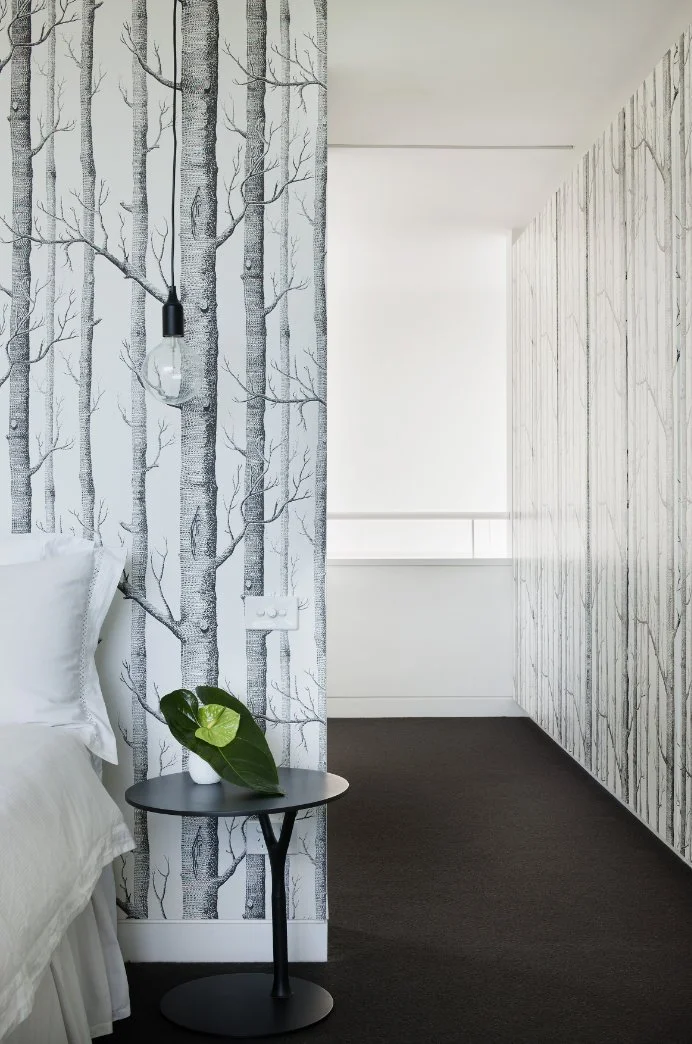 Schlafzimmer mit weißem Bett und schwarzem Beistelltisch vor einer schwarz-weißen Baumtapete.