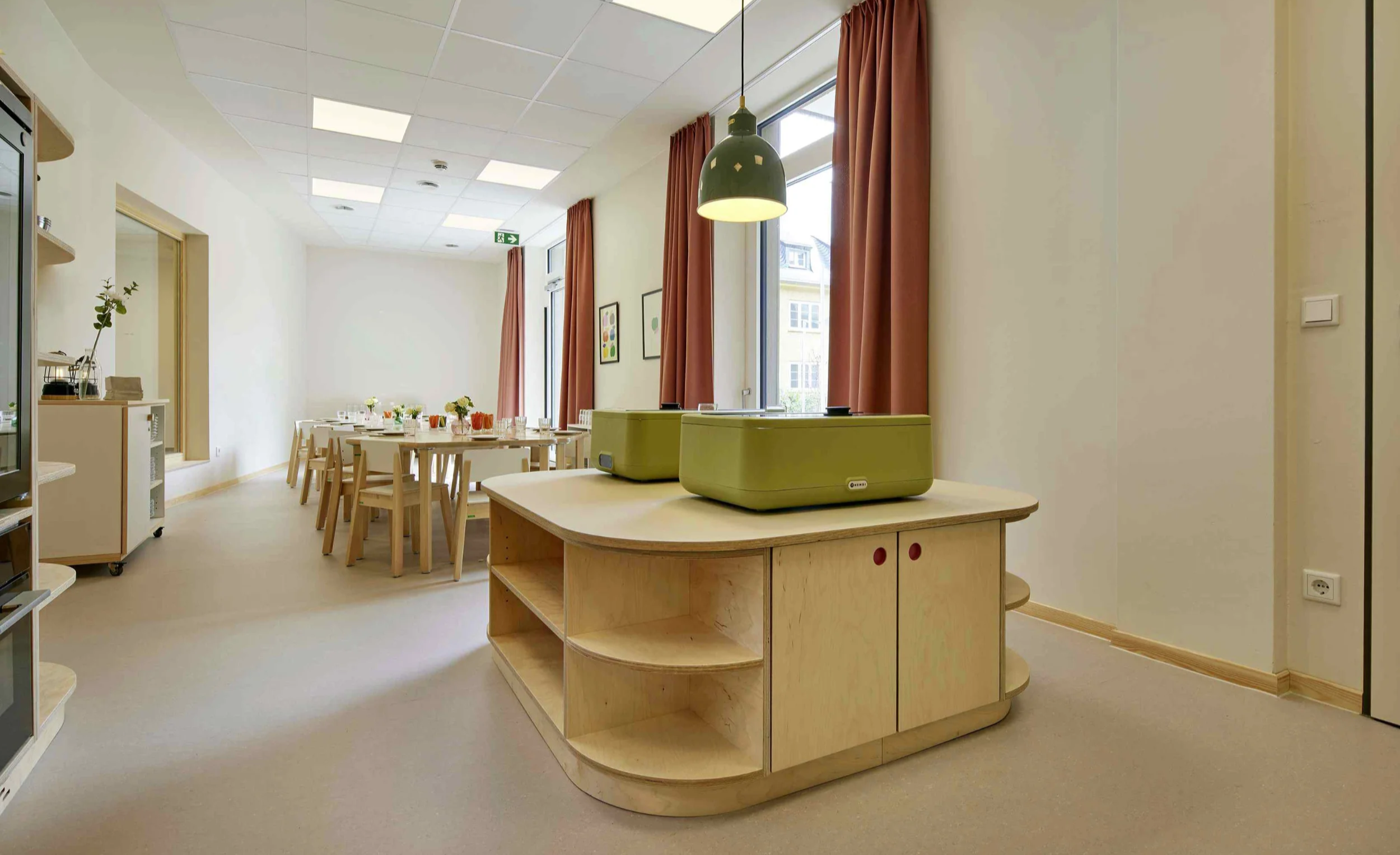 Ein heller Raum mit einem Holztheken, auf dem zwei grüne Geräte stehen. Im Hintergrund ein Esstisch mit Stühlen und gedecktem Tisch, vor Fenstern mit roten Vorhängen. An der Wand hängen einige Bilder.