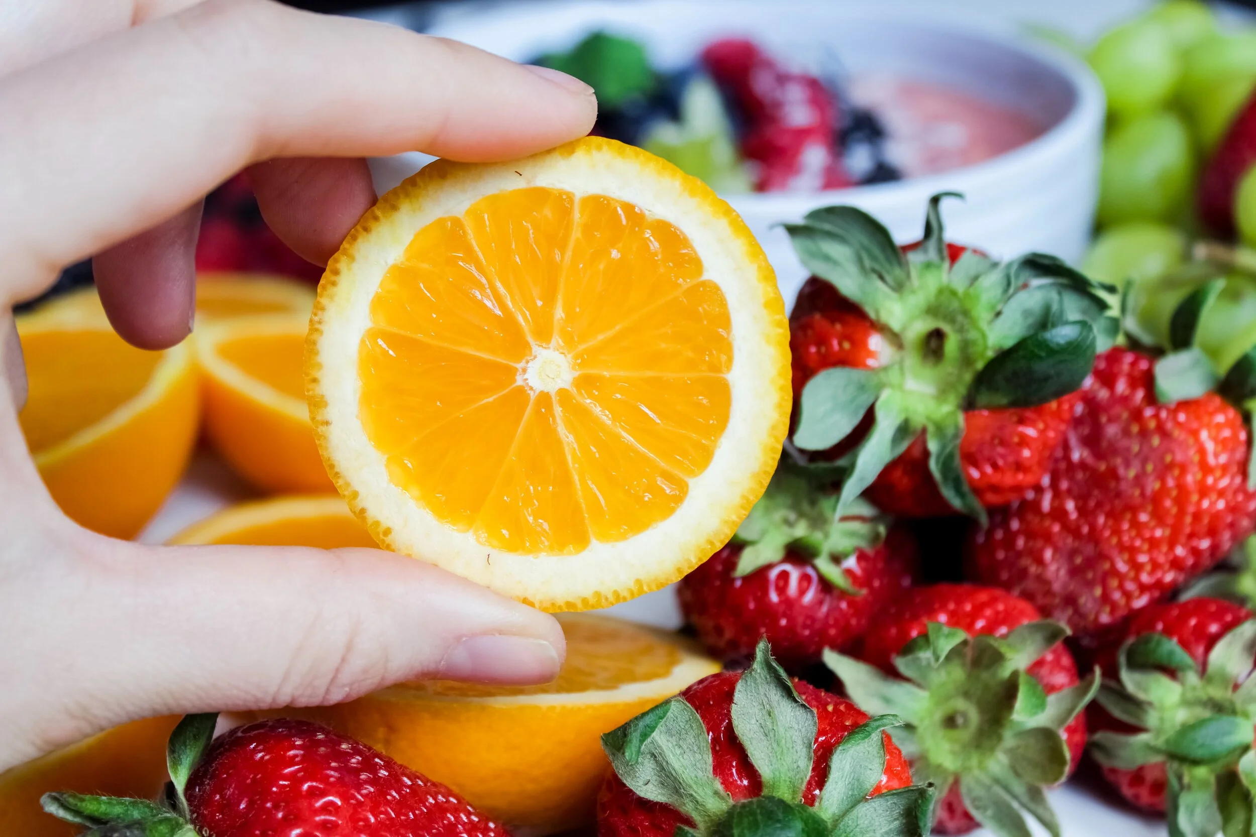 Hand holding half of an orange with a background of strawberries and other sliced oranges and berries.