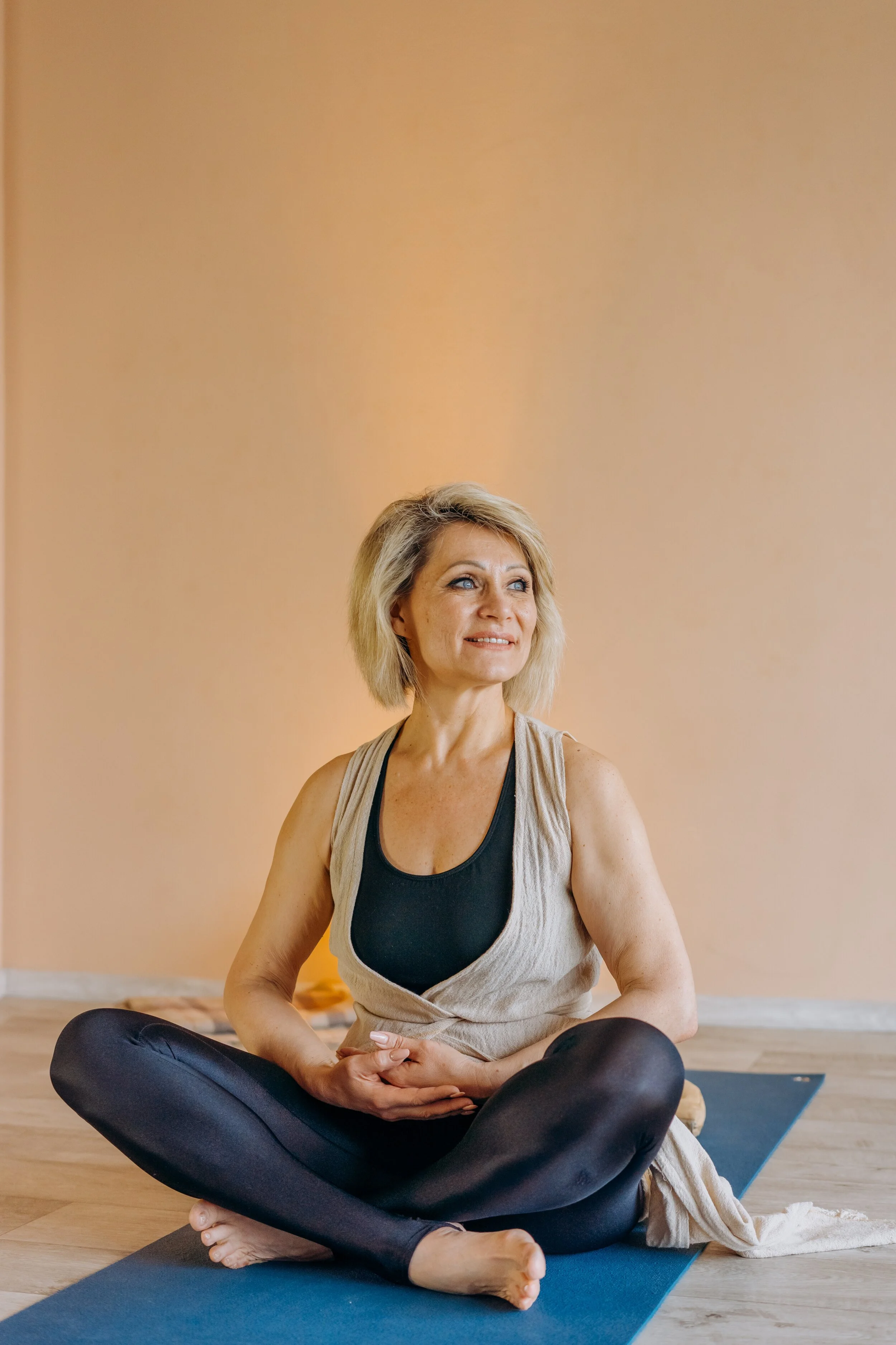 A middle-aged woman with blonde hair practicing yoga indoors on a blue mat, sitting cross-legged with hands resting on her stomach, in front of a plain beige wall.