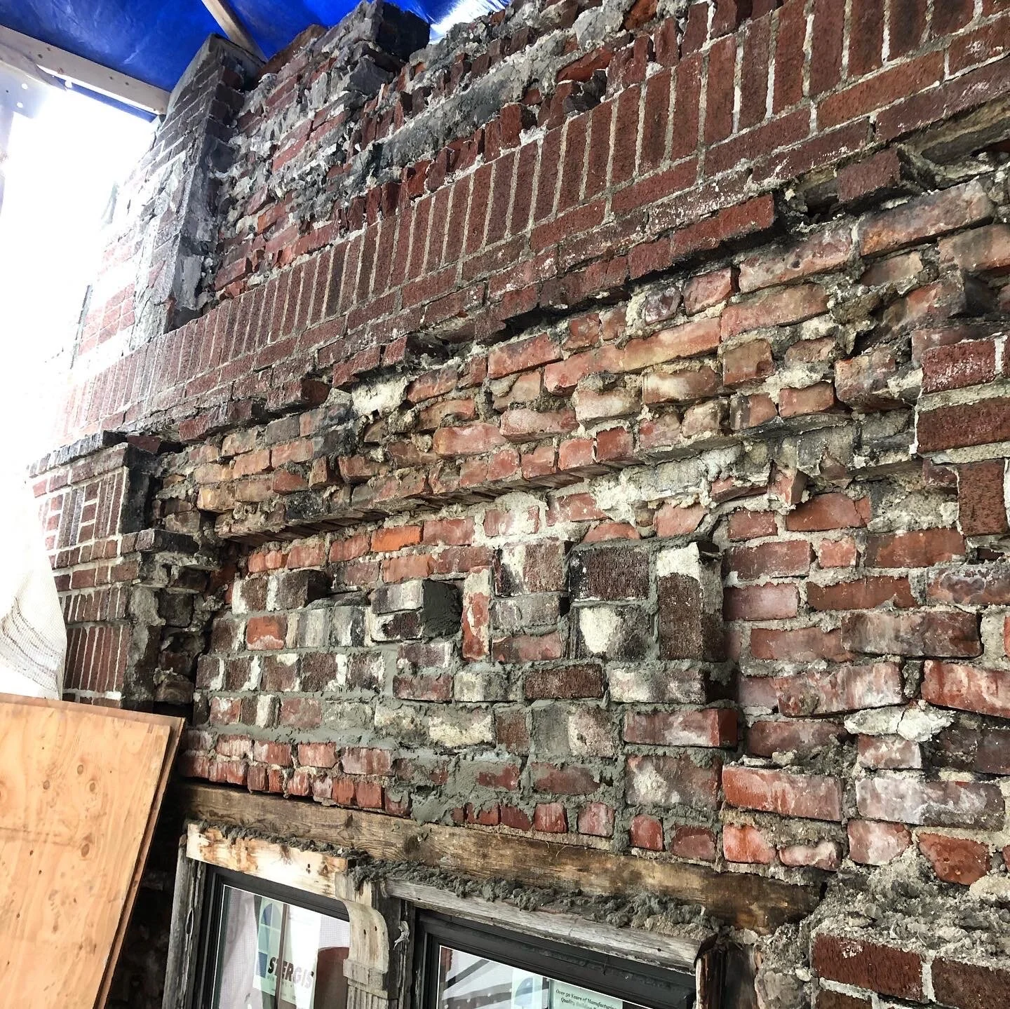 Close-up of a deteriorating brick wall with exposed mortar and bricks in various stages of decay. A blue tarp partially covers the structure above, and there is a wooden frame at the bottom.
