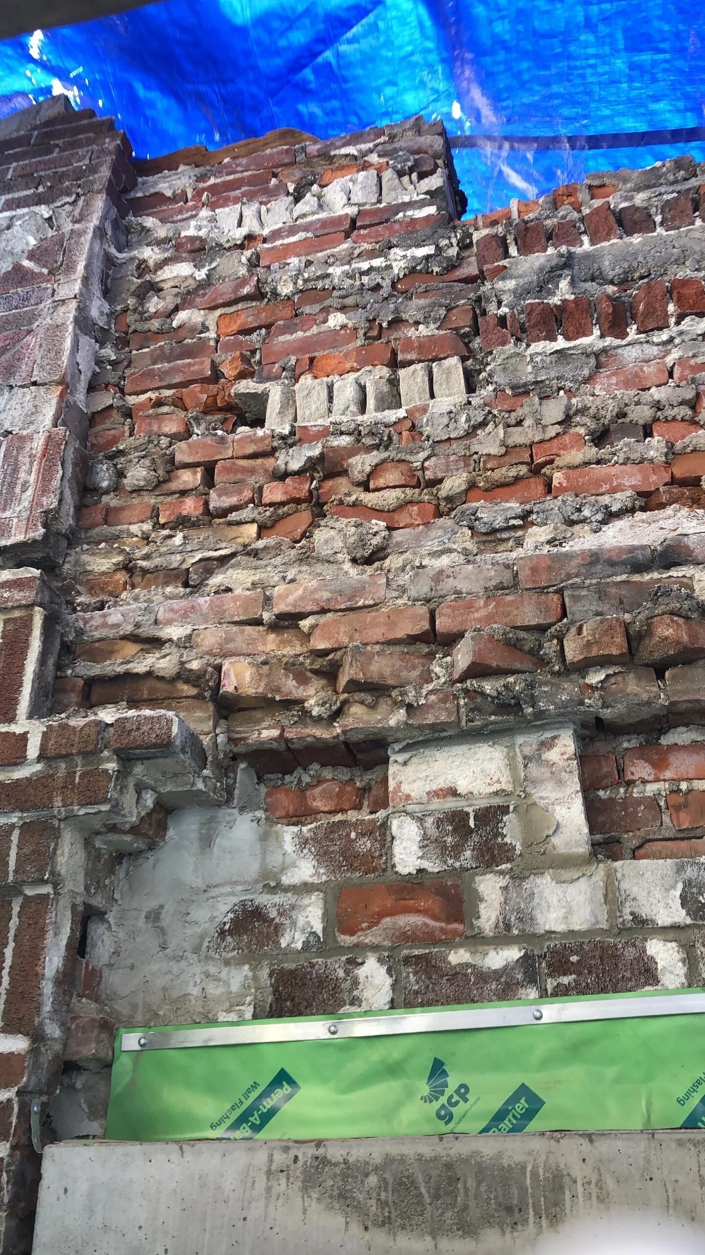 Old brick wall with visible deterioration covered by a blue tarp, showing crumbling mortar and some newer construction materials at the base.