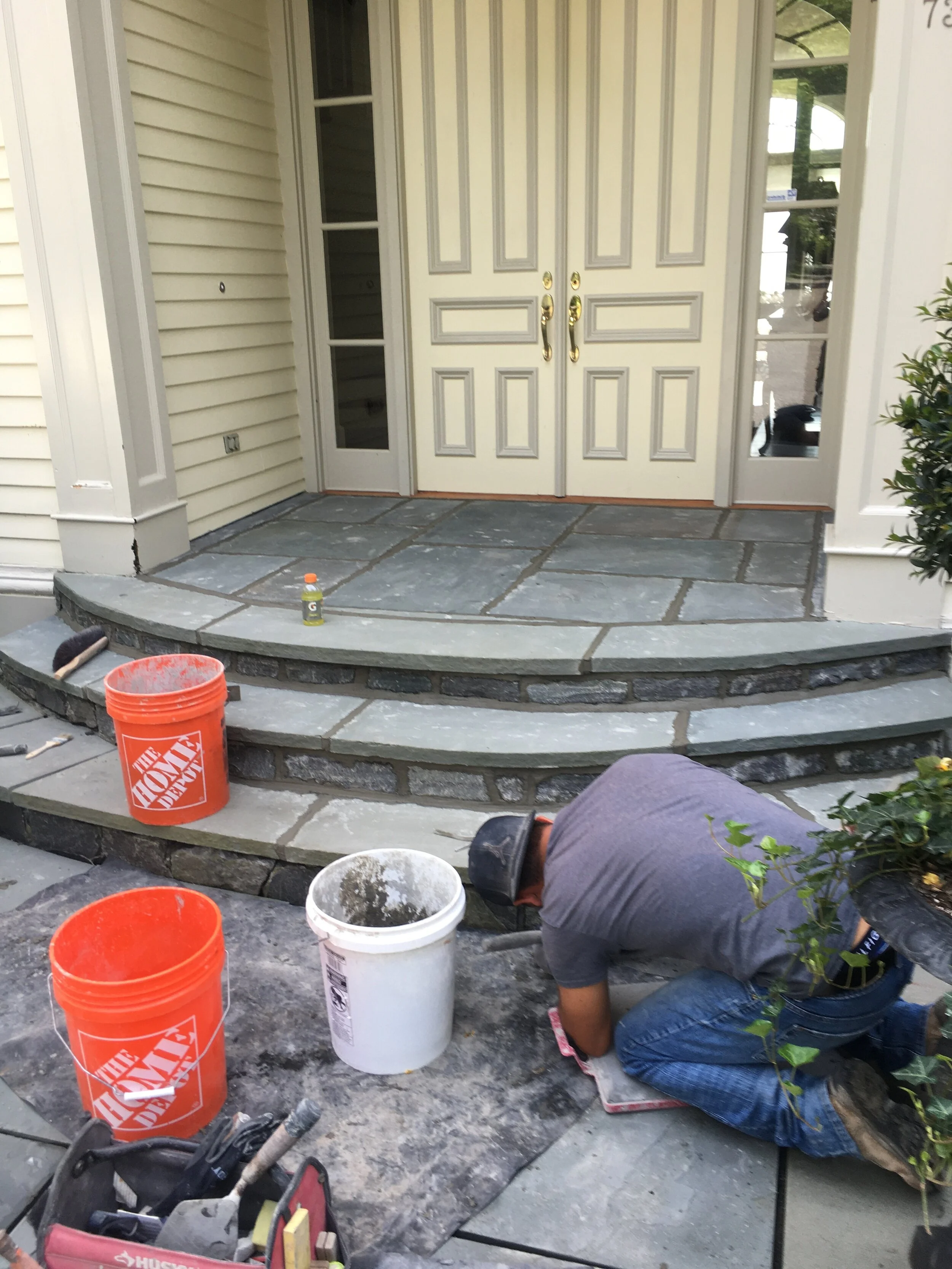 Construction worker laying stone tiles on a front porch with buckets and tools nearby.