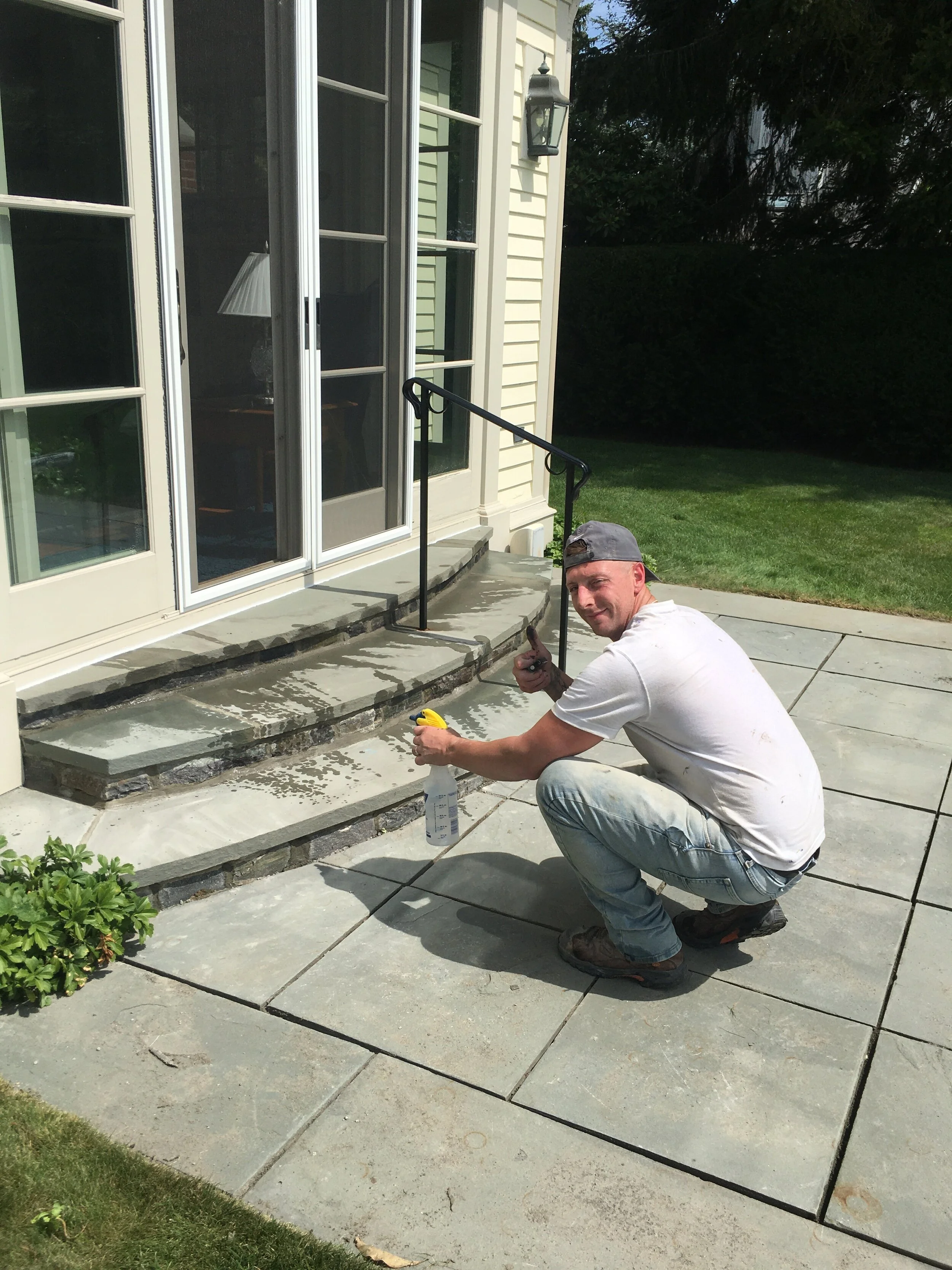 A person in a white t-shirt and jeans is crouching on a stone patio, working on a set of outdoor steps with a spray bottle and trowel. The steps lead up to a set of glass doors on a light-colored building. A metal handrail is installed on the steps, 