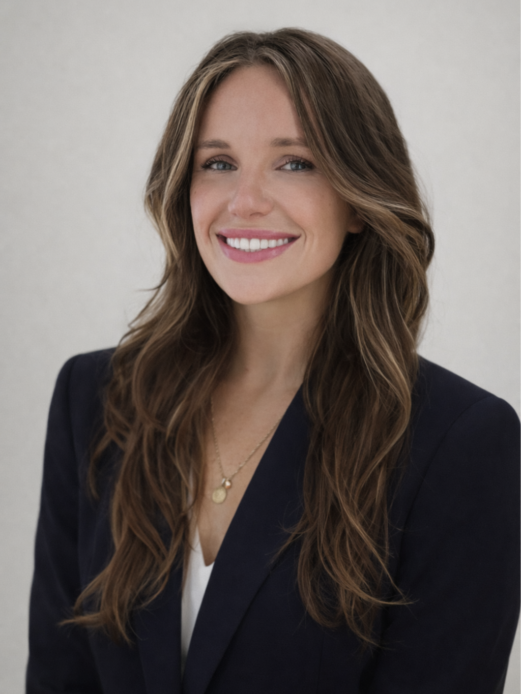 A woman with long brown hair styled in loose waves, smiling, wearing a black blazer and a white top, with a gold necklace, standing against a plain light-colored background.