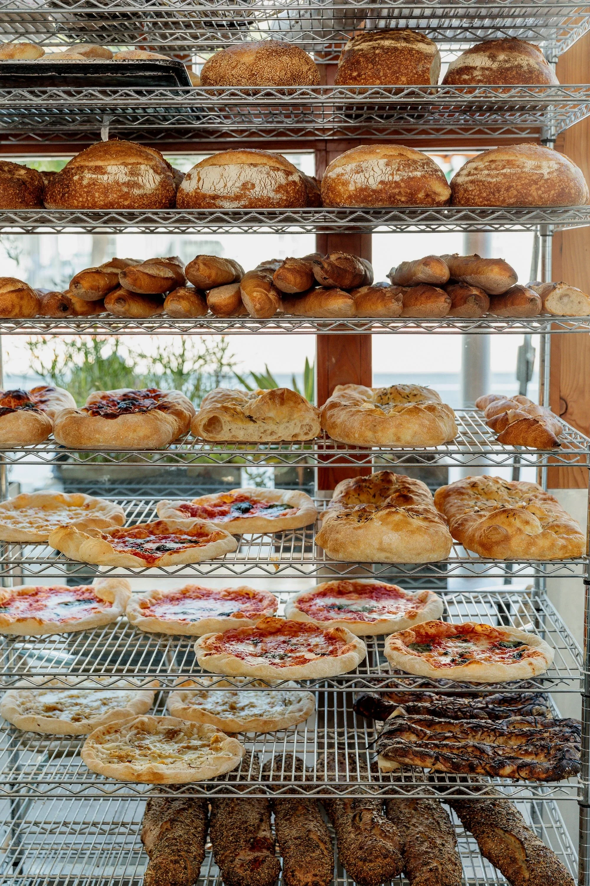 Photo of bakery cooling rack stacked with fresh baked goods like pizzas and breads.