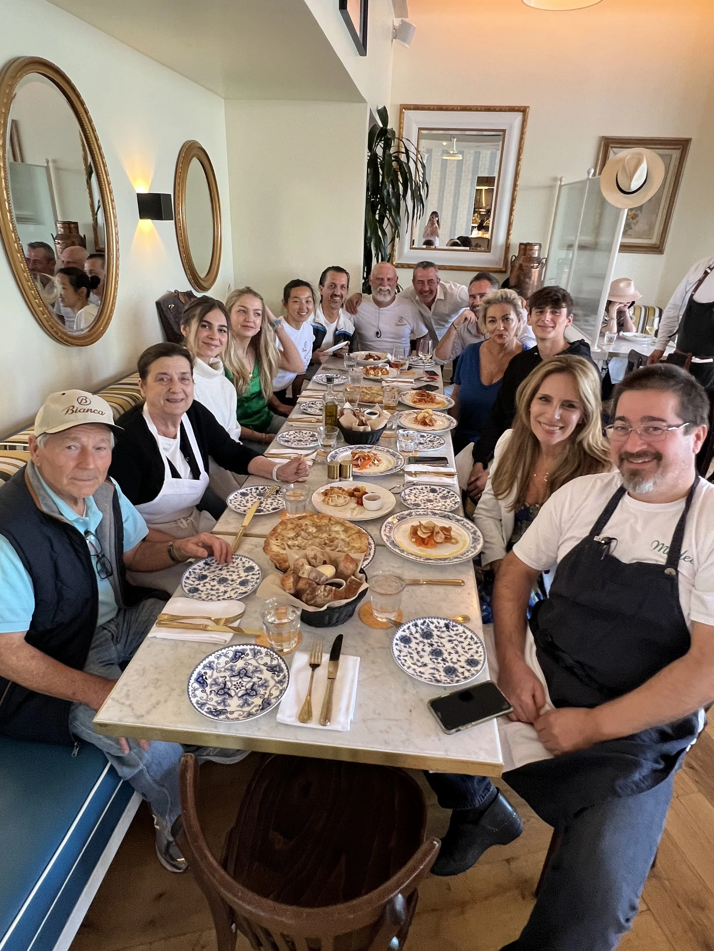 A group of people sitting around a table in a restaurant, smiling for a group photo. The table is set with plates, cutlery, and a variety of food dishes. The restaurant decor includes mirrors and framed pictures on the walls.