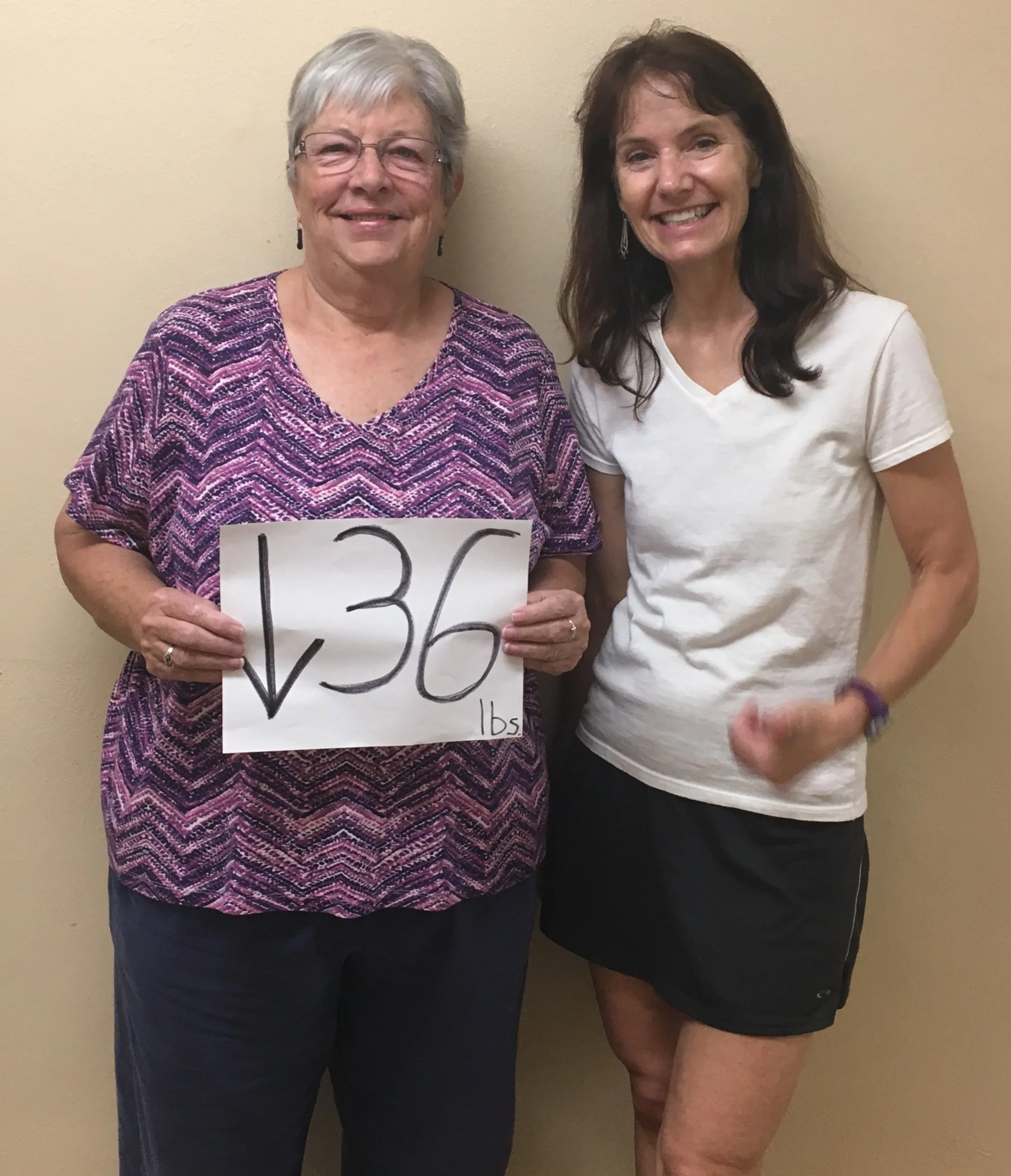 Coach Gayle stands with her health and fitness coaching client, Mary Ellen, who is holding a sign with a down arrow and the number 36 to indicate her weight loss success. Women's whole health and certified fitness coach.