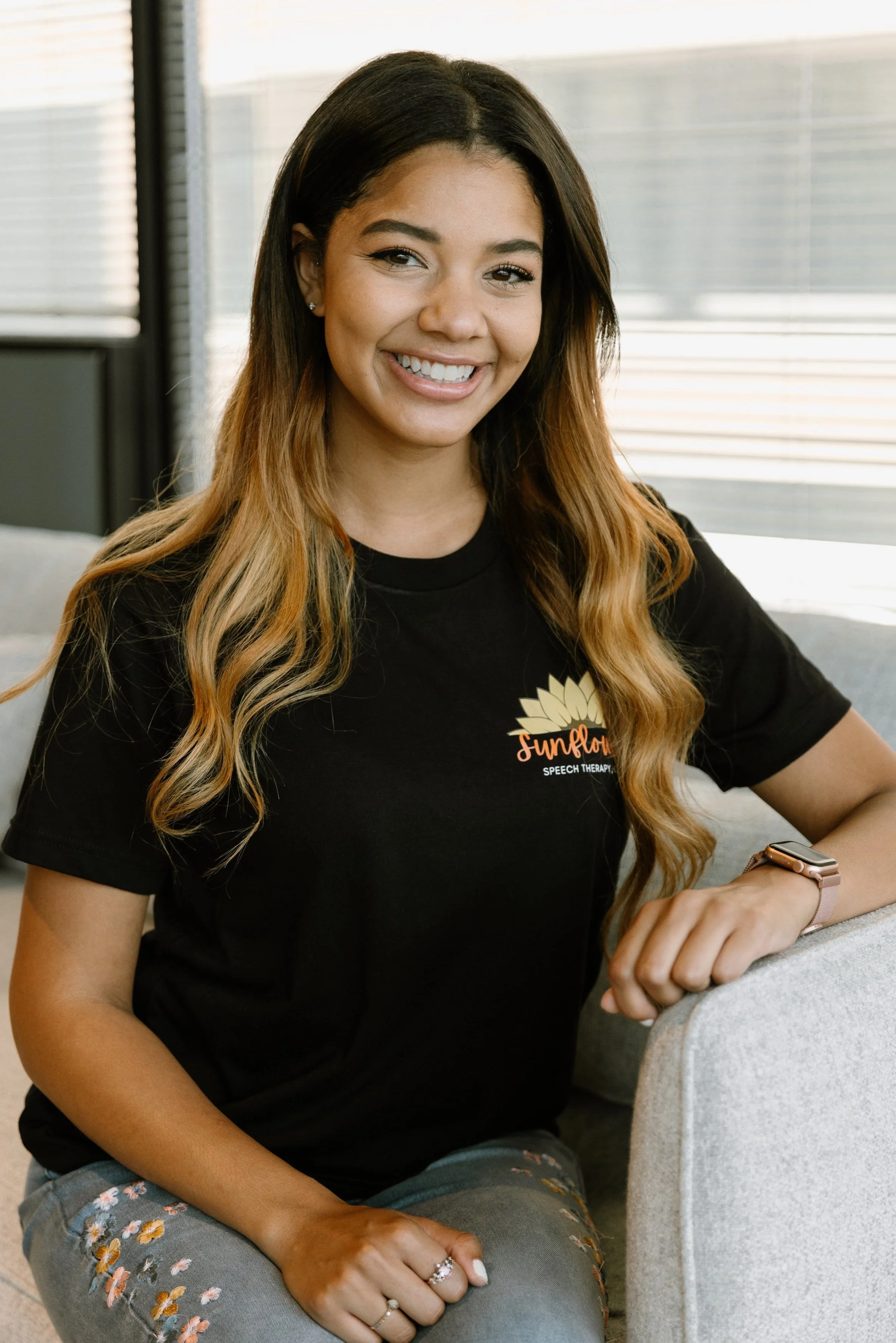A smiling woman with long, wavy hair wearing a black t-shirt with a sunflower logo and the words "Sunflower Speech Therapy." She is sitting on a gray couch, with her left elbow resting on the armrest and her right hand on her lap, in a well-lit room with blinds in the background.