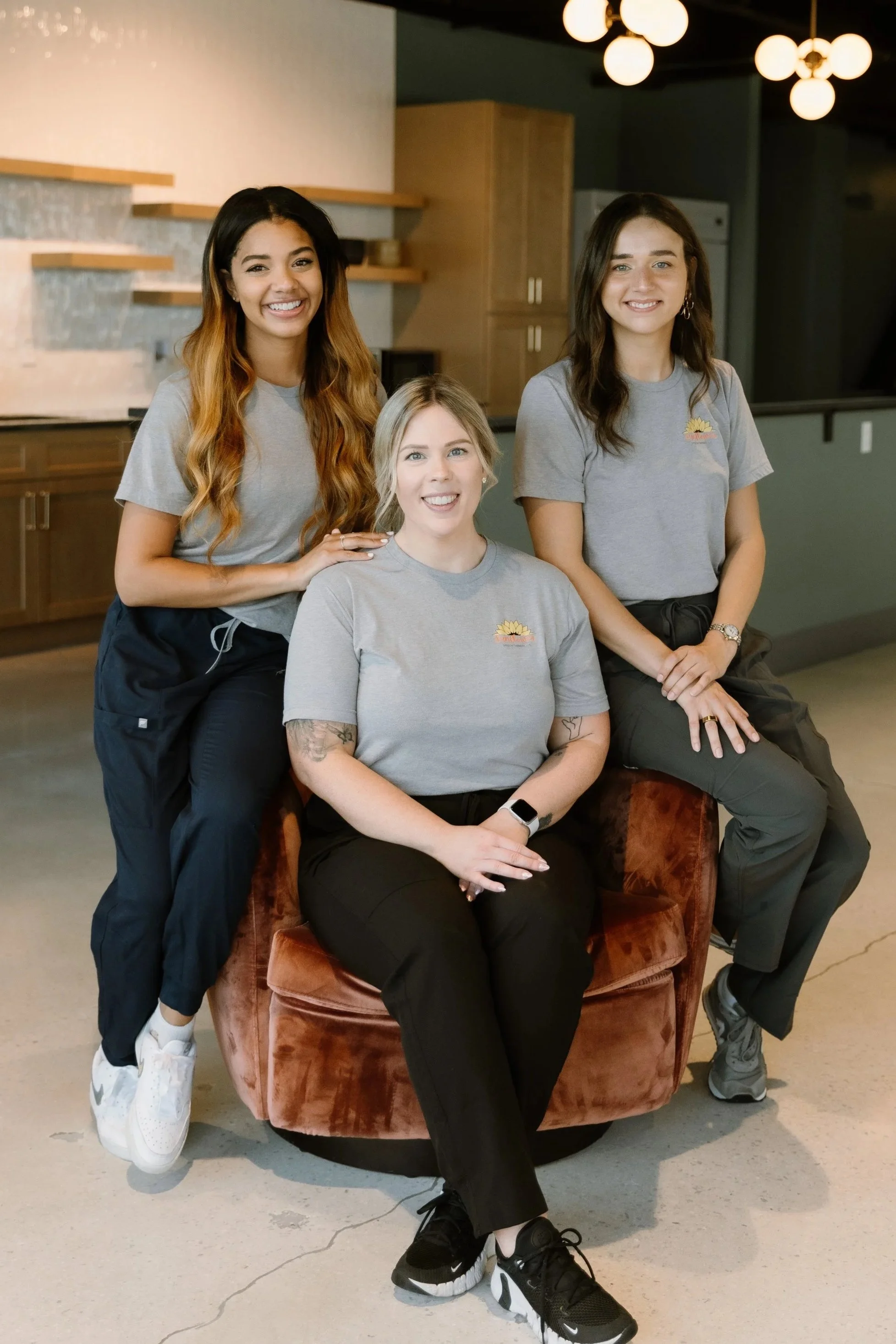 Three women in gray t-shirts and black pants, two standing and one sitting on a swivel chair, smiling in a modern indoor space with warm lighting.