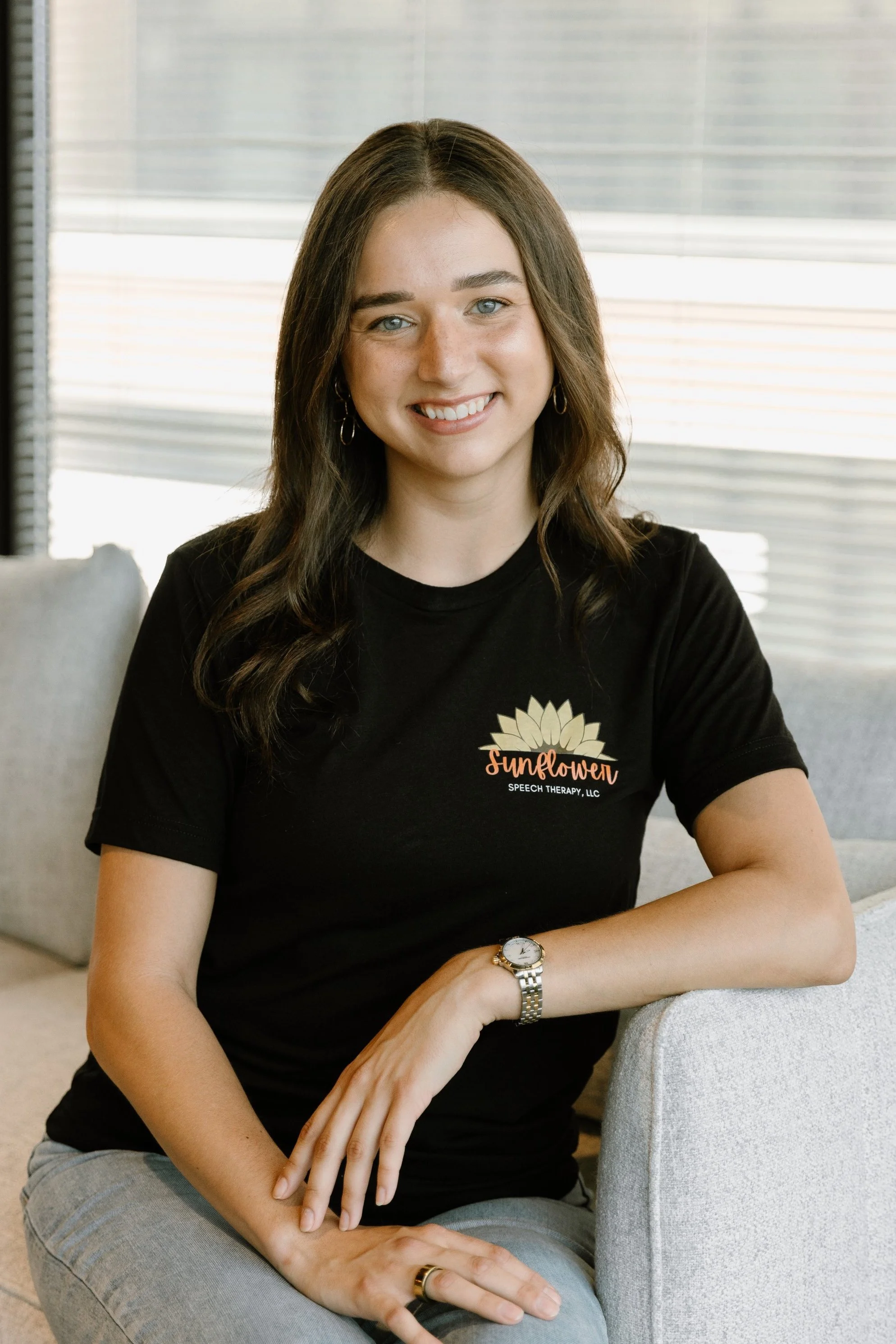 A young woman with shoulder-length brown hair, blue eyes, and a smile, sitting on a light-colored sofa in a bright room. She is wearing a black t-shirt with a sunflower logo and the words 'Sunflower Speech Therapy, LLC,' along with a silver watch, a ring, and hoop earrings.