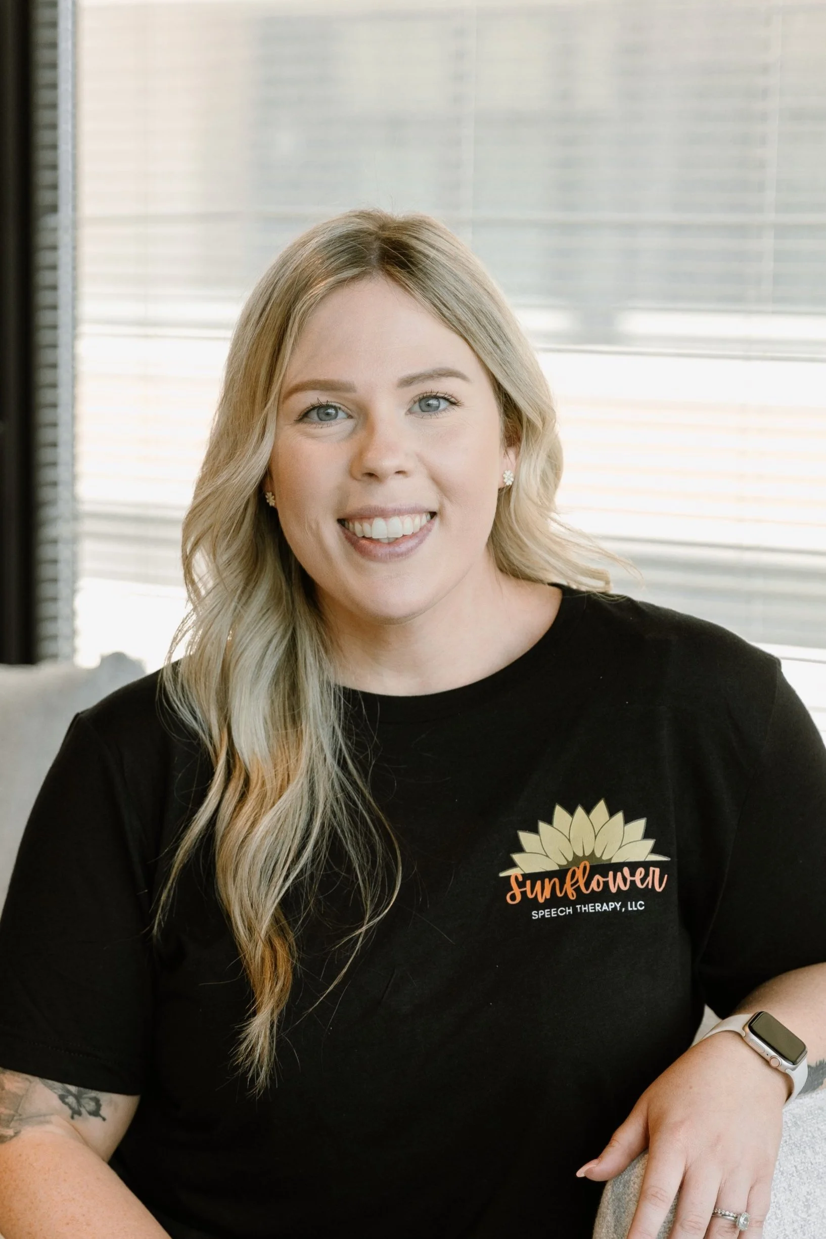 A woman with blonde hair and blue eyes, smiling, wearing a black t-shirt with a sunflower logo and the words 'Sunflower Speech Therapy, LLC.' sitting indoors with window blinds in the background.