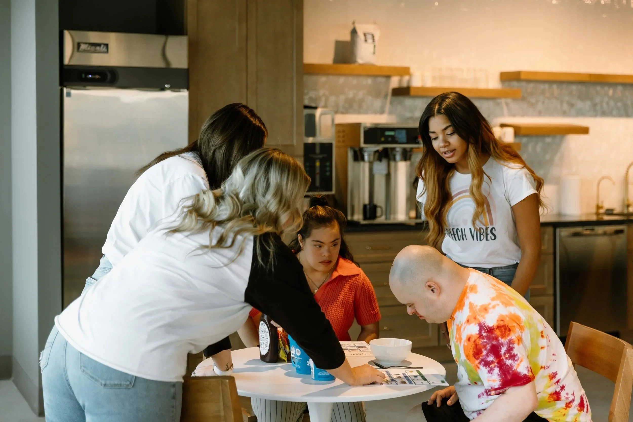 Five people are gathered around a table in a kitchen, looking at a low tech communication board. One person is seated, another is leaning over, and three others are standing around, observing and discussing.
