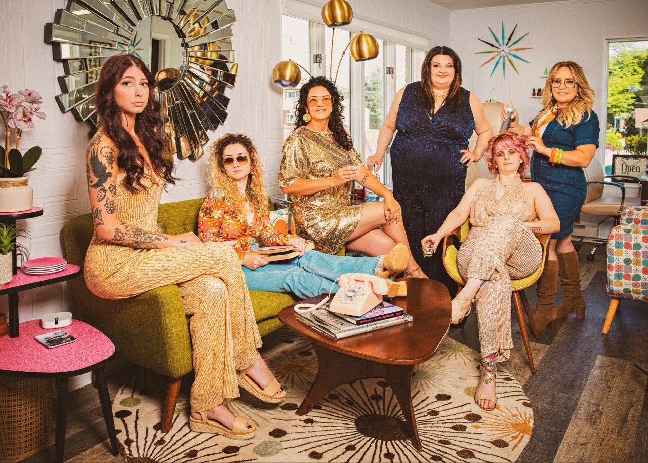 A group of six women in a brightly decorated living room, sitting and standing around a coffee table with a vintage rotary phone.