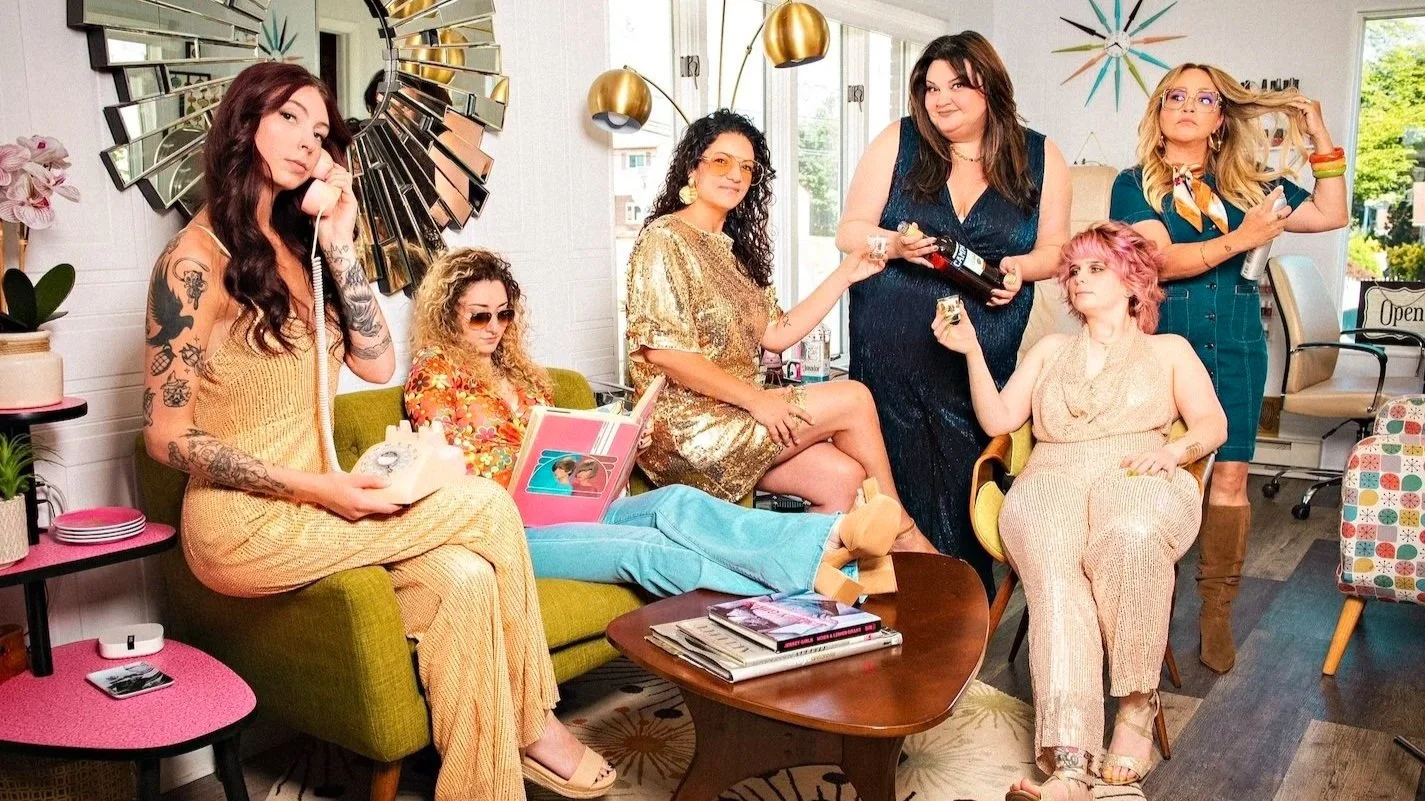 Six women in vintage clothing in a retro-style living room with colorful decor, some holding drinks or phones, various poses and expressions.