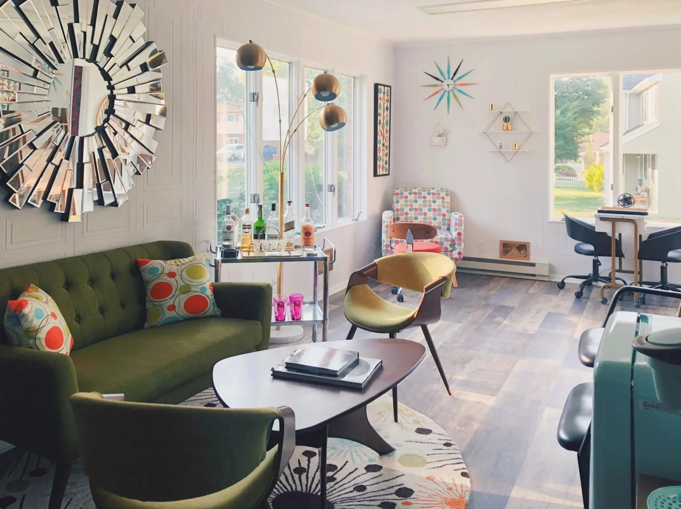 A bright living room with white walls and large windows letting in natural light. There is a green sofa with colorful polka dot cushions, a modern armchair with a wood back, and a coffee table with stacked books. Decorative elements include a large sunburst mirror, a multi-armed floor lamp with metallic globes, wall art, geometric shelves with small plants and items, and a patterned area rug.