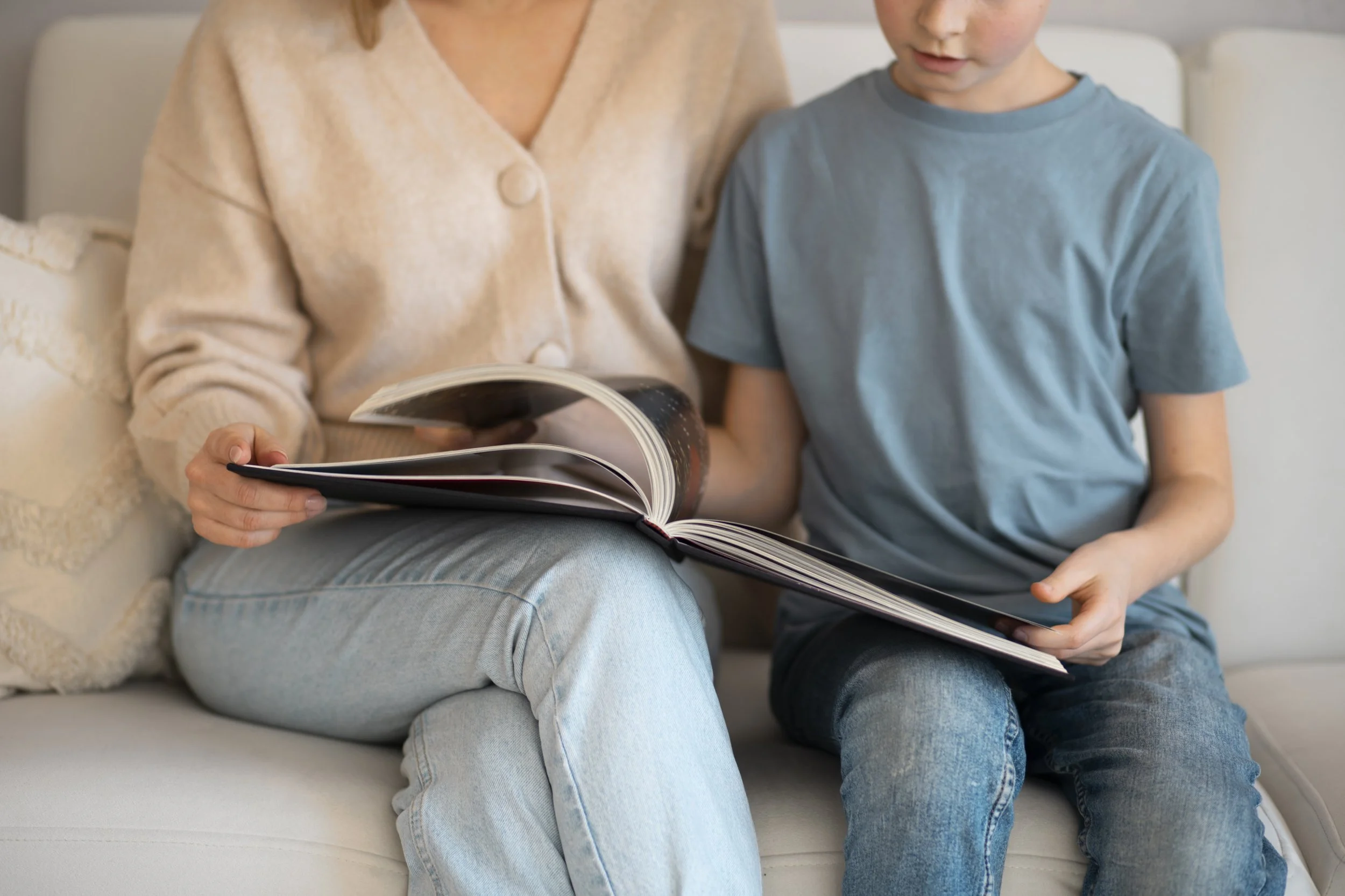 A woman and a young boy sitting on a beige sofa, looking at a magazine together.
