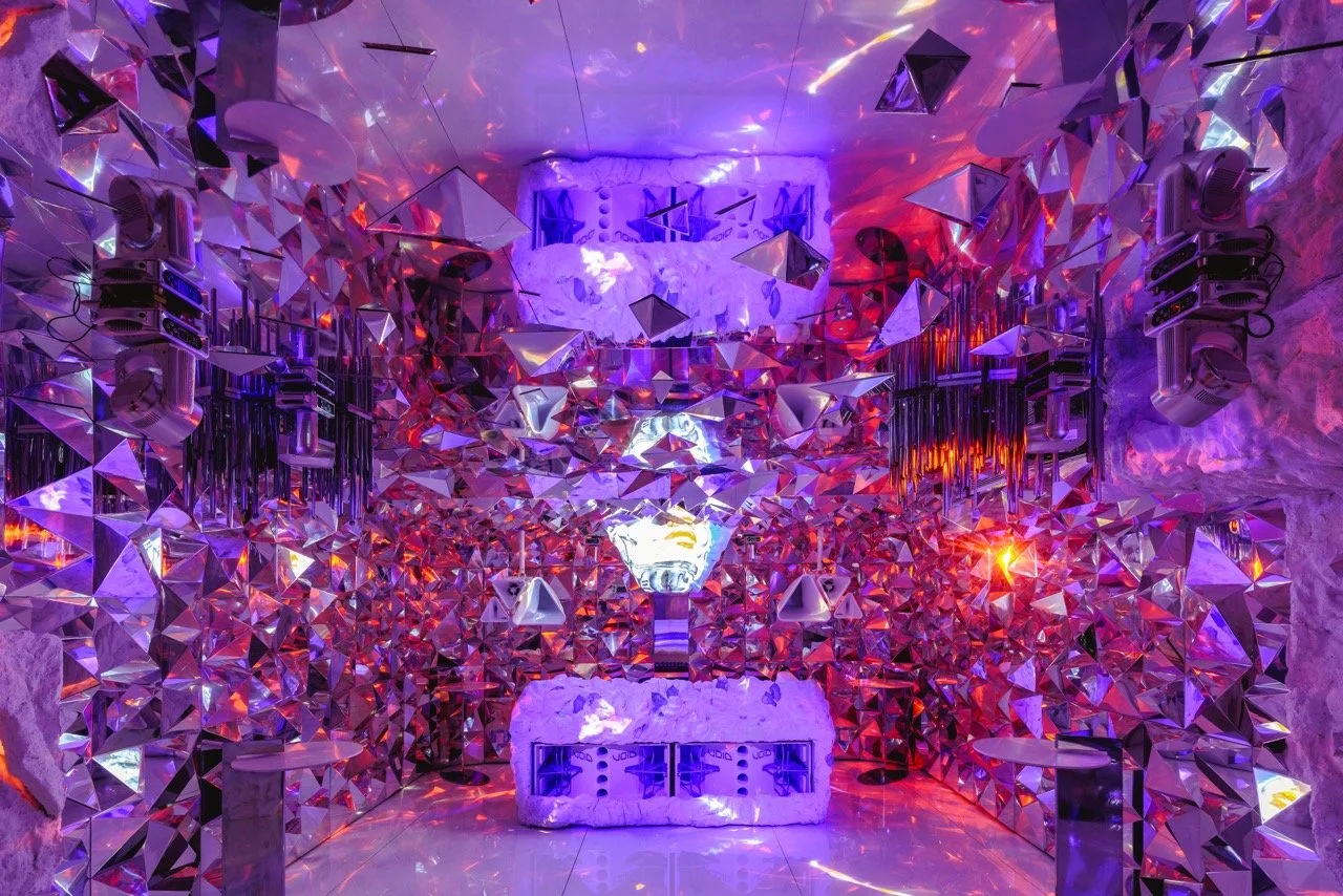 Interior con paredes cubiertas de espejos y cristales en forma de triángulos, iluminación en tonos violetas, azules y rojos, con luz en el centro y objetos decorativos en la parte superior, ambiente futurista y brillante.