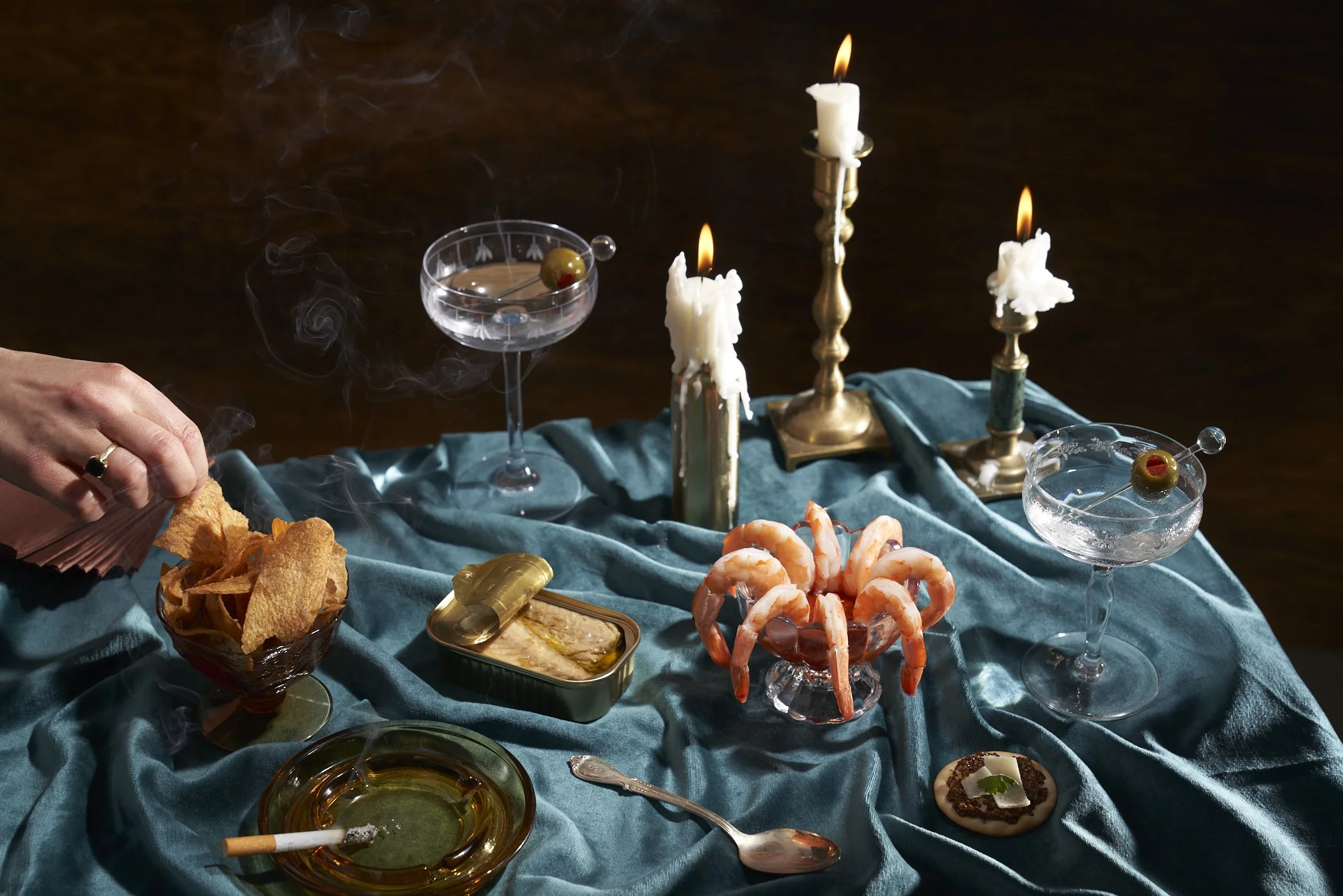 A moody dinner party scene with candles, surrounded by shrimp, cigarettes, tin fish and martini cocktails. 