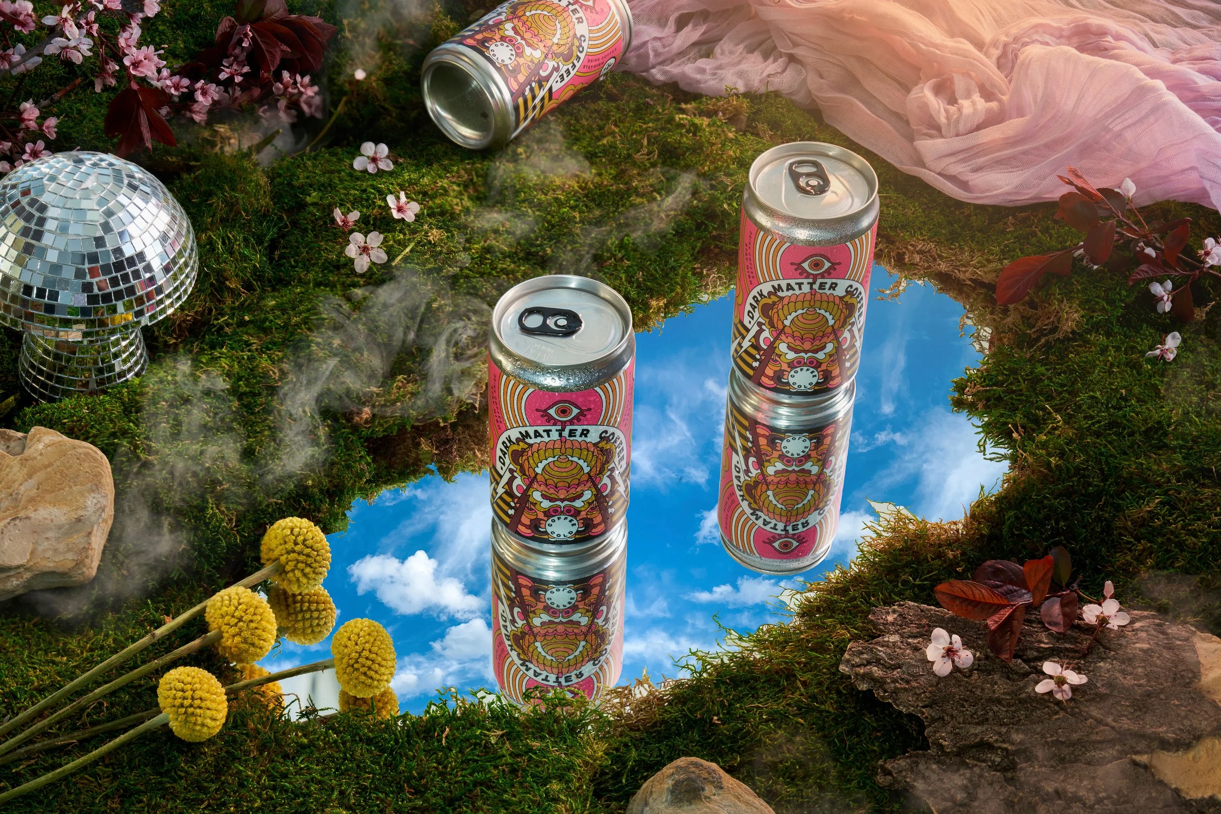 Two cans of Dark matter iced coffee in a can.  Sitting on a pool of water with a refection of the sky. There is a green moss atmosphere around it with a soft pink blanket, disco ball mushroom and natural elements.  Some fog rolls off the grass.