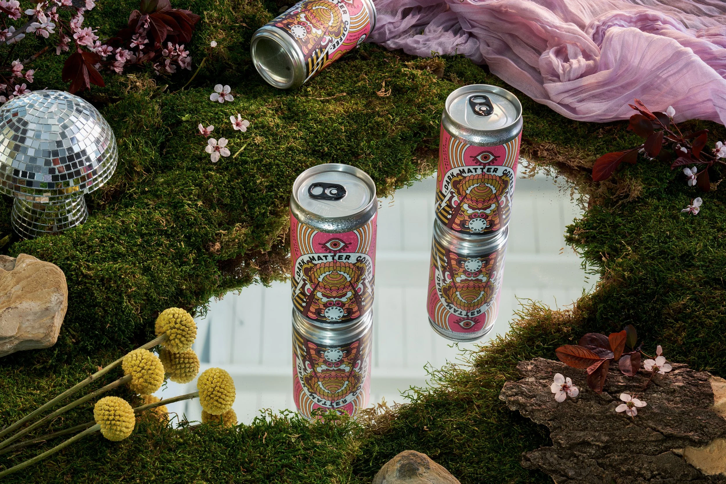 Two cans of dark matter coffee sitting on a mirror surrounded by moss and natural elements