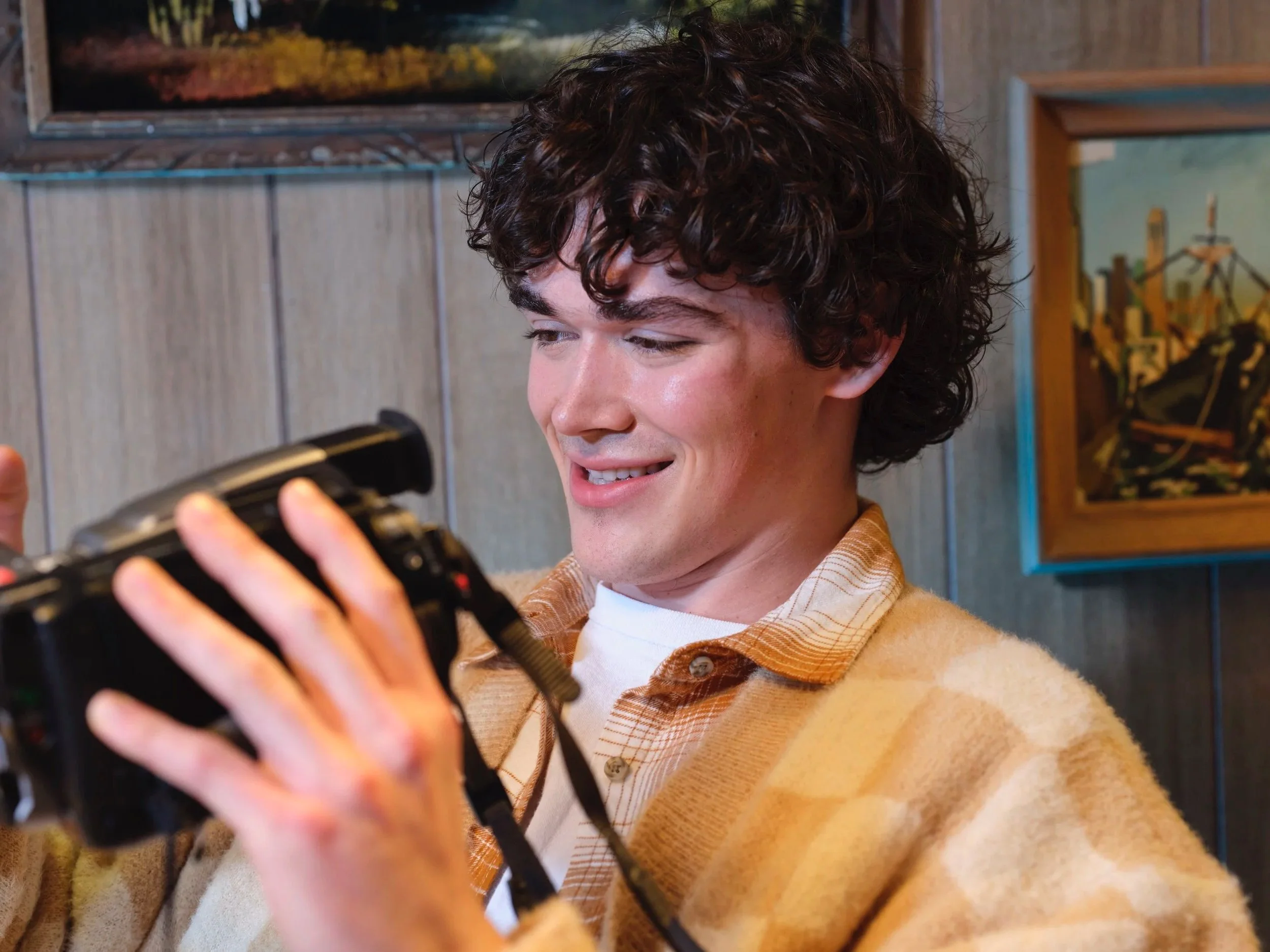 Young man with curly brown hair smiling while holding a camera indoors, with framed pictures hanging on a wooden wall behind him.