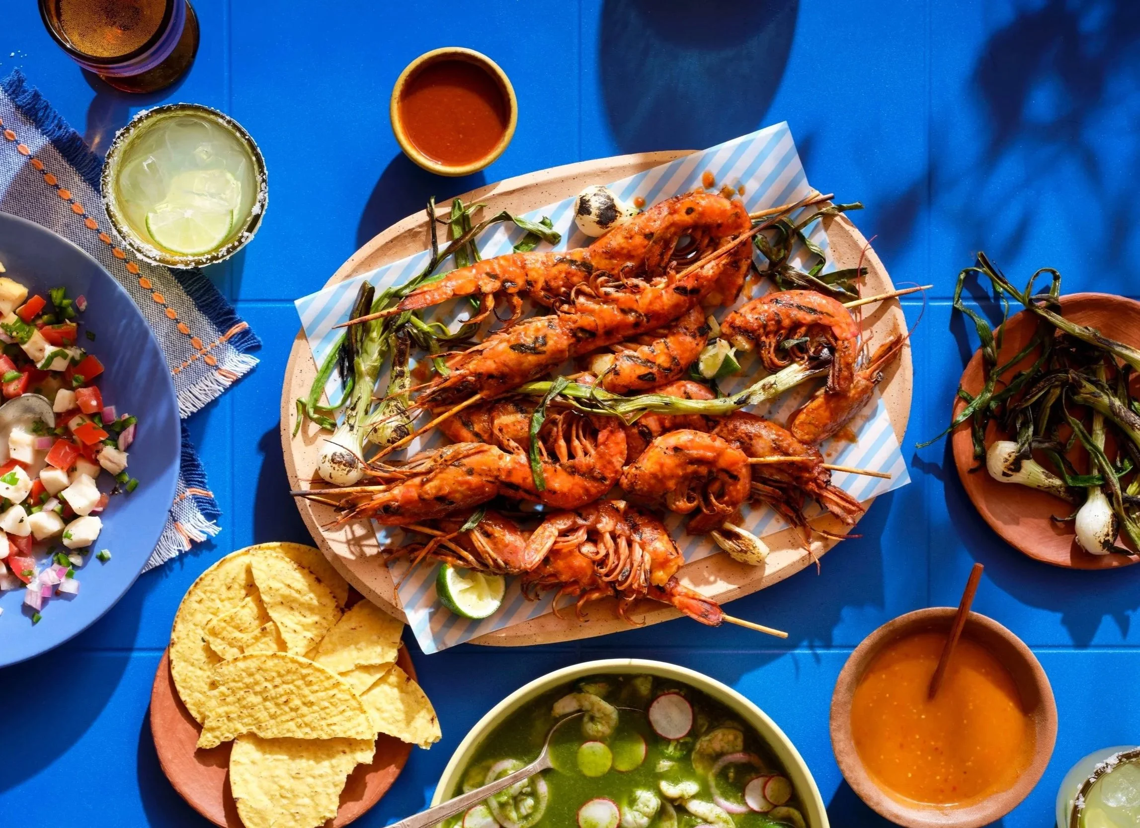 A blue tile table with warm sunlight and dark shadows.  On the table is a platter or shrimp, grilled green onions, salsa, ceviche, margaritas and tostadas.