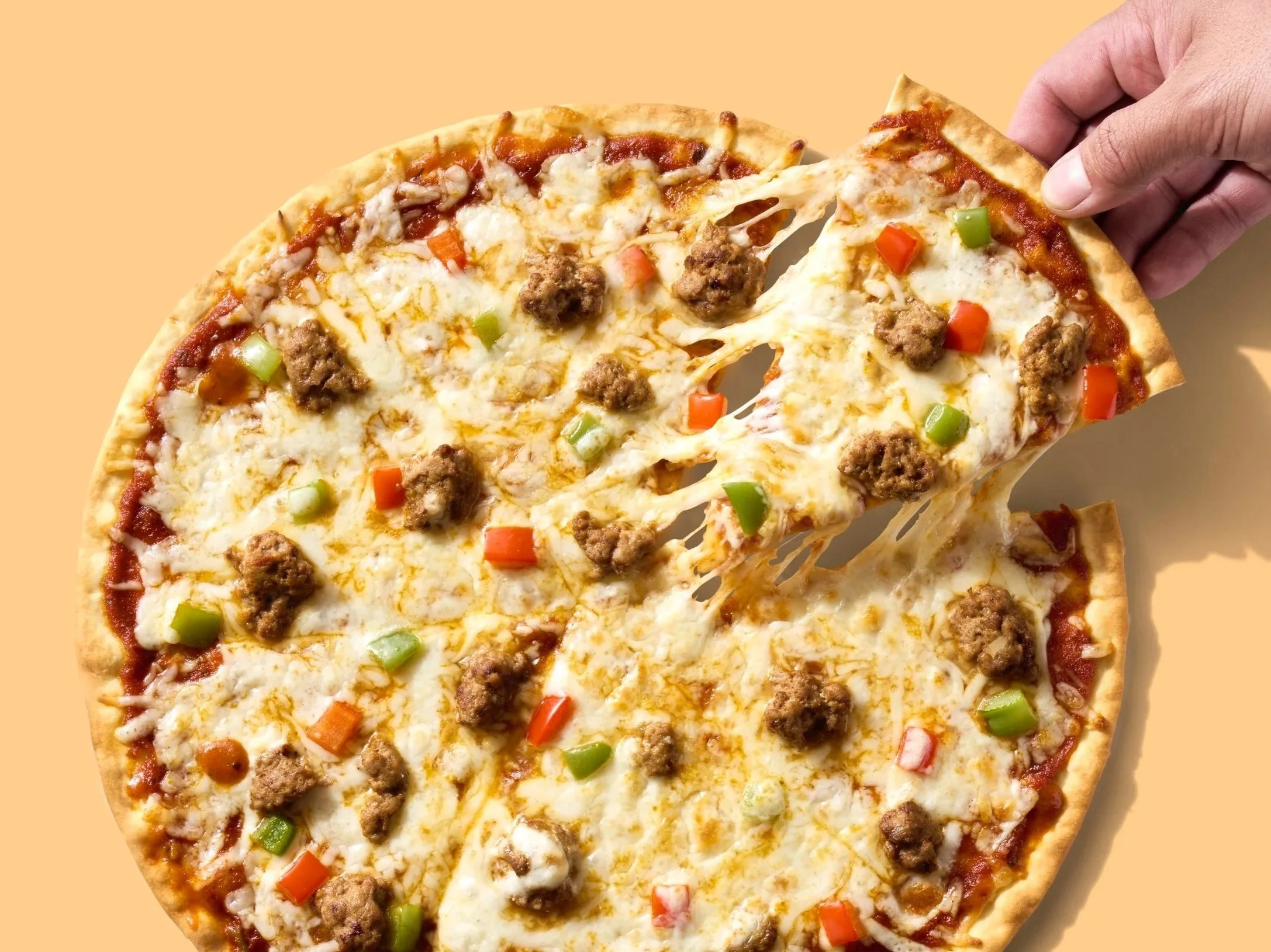 A hand pulling a slice of pizza from a whole pie. It is sitting on a yellow orange background. 
