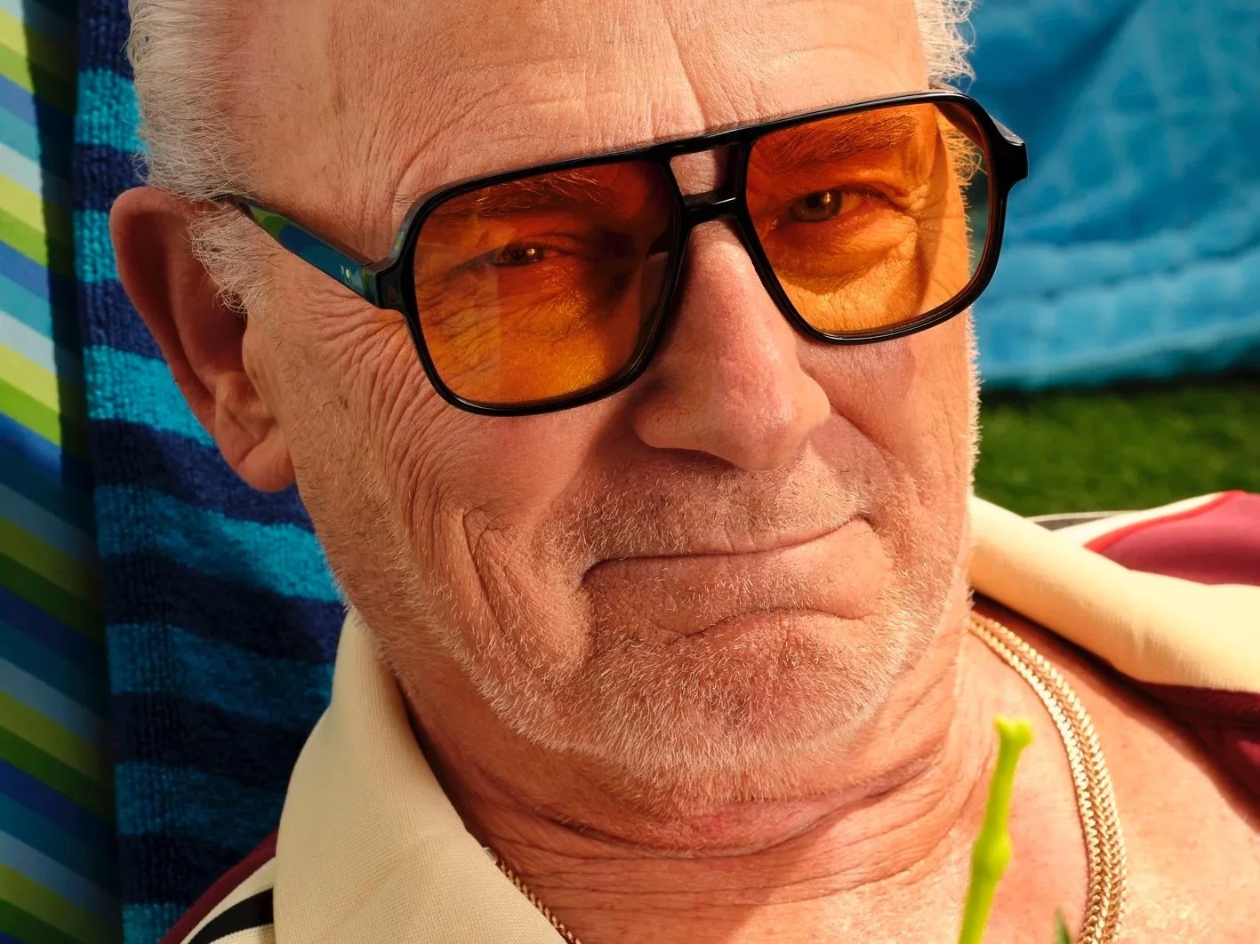 man sitting on a beach chair in a speedo and track jacket surrounded by a kiddie pool, radio, beer, charcoal grill and sun tan oil