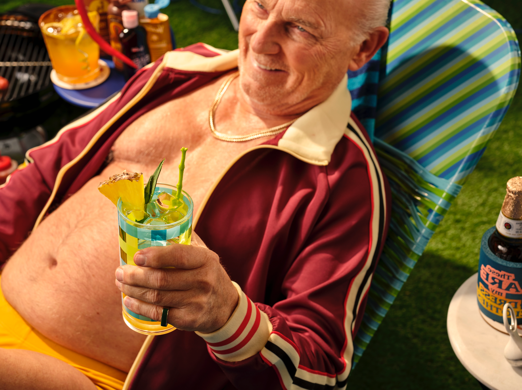 man sitting on a beach chair in a speedo and track jacket surrounded by a kiddie pool, radio, beer, charcoal grill and sun tan oil