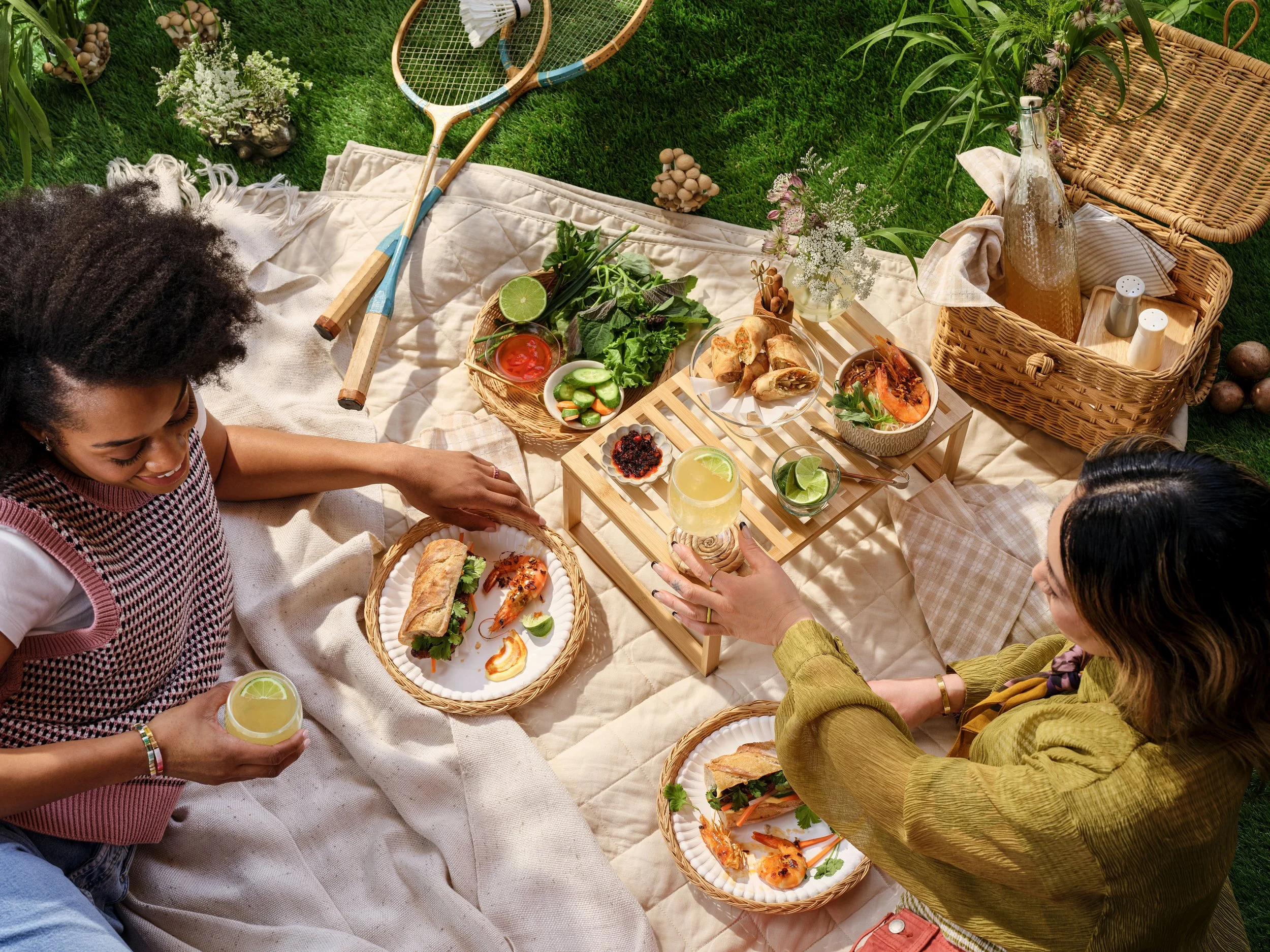 two women laying on a picnic blanker in the park, sharing food and drink on a summer day
