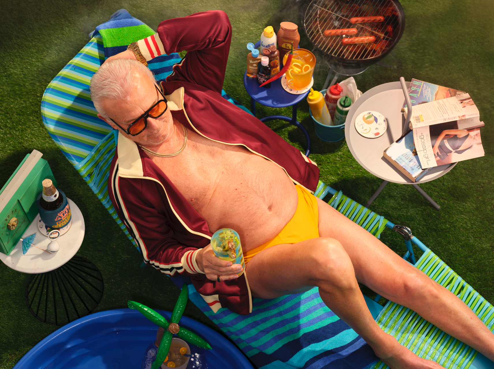 man sitting on a beach chair in a speedo and track jacket surrounded by a kiddie pool, radio, beer, charcoal grill and sun tan oil