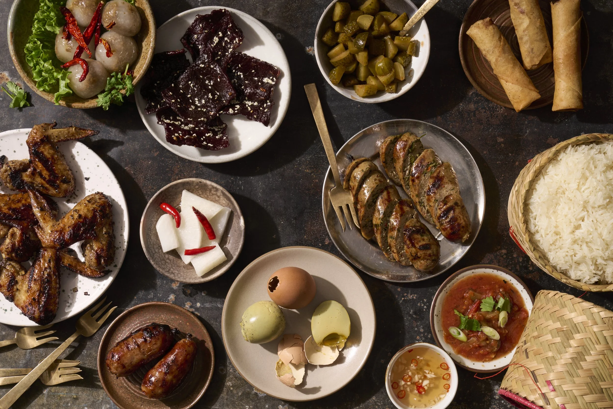 Assorted Vietnamese dishes on a dark table, including spring rolls, grilled meat, boiled chicken wings, pickled vegetables, boiled eggs, rice, dipping sauces, and a small basket.