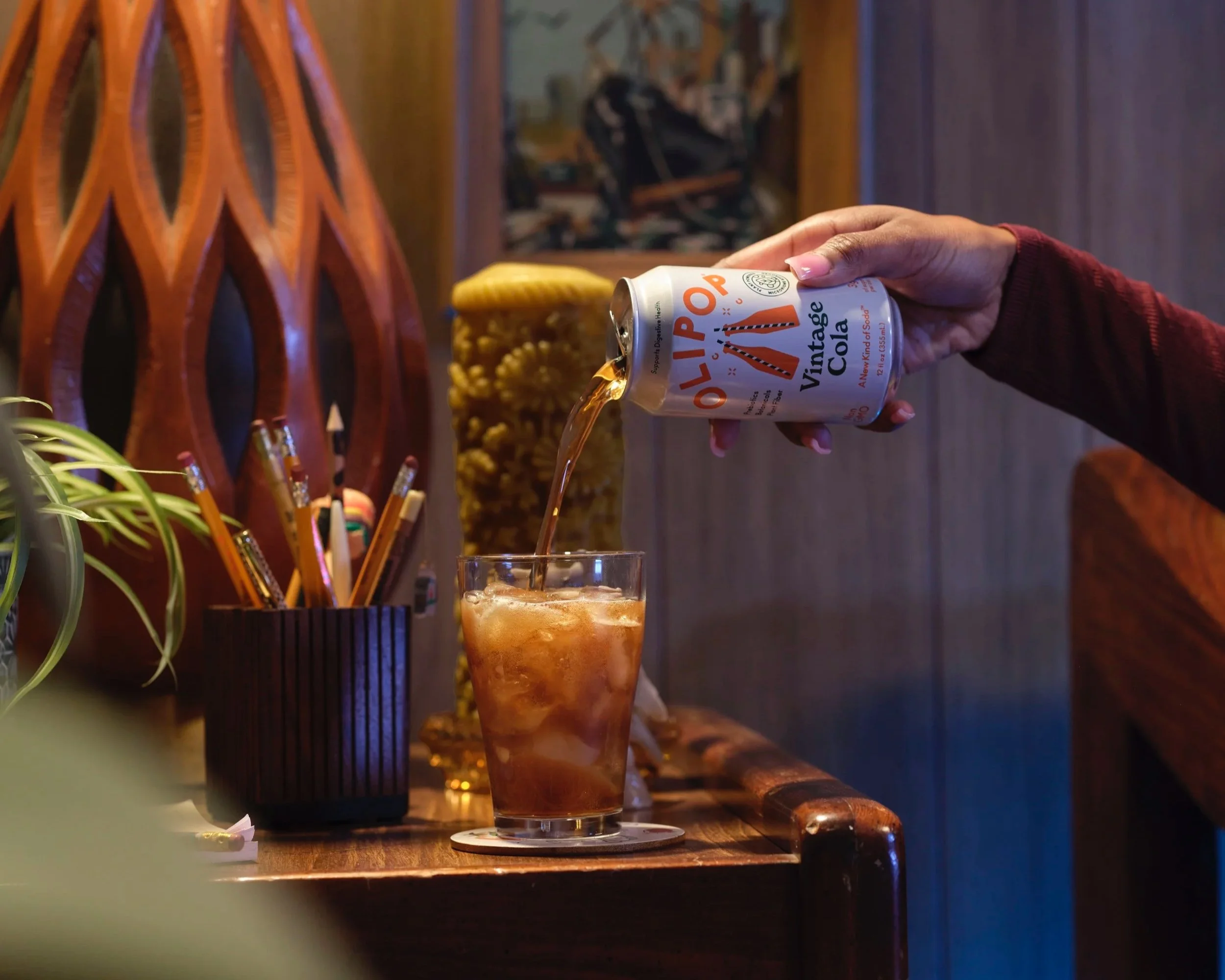 Can of olipop vintage cola being poured into a glass with ice.  The glass is sitting on a wooden side table with a container of pens and pencils. The hand holding the can is a ladies hand with pink nails.