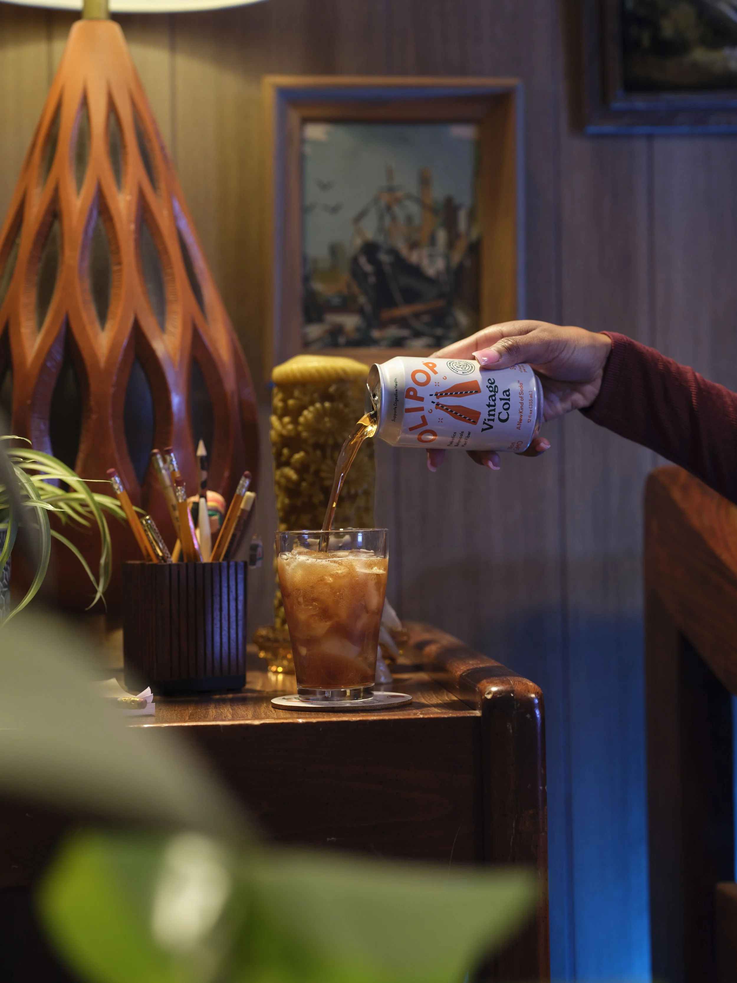 A person pouring OyliPOP Vintage Cola from a can into a glass of ice on a wooden table surrounded by decorative items and paintings in a cozy room.