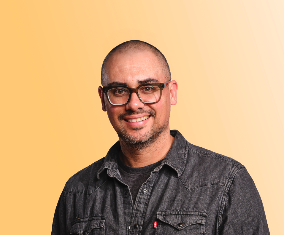 Man wearing a black denim shirt and green glasses, short black hair and black and grey facial hair.
