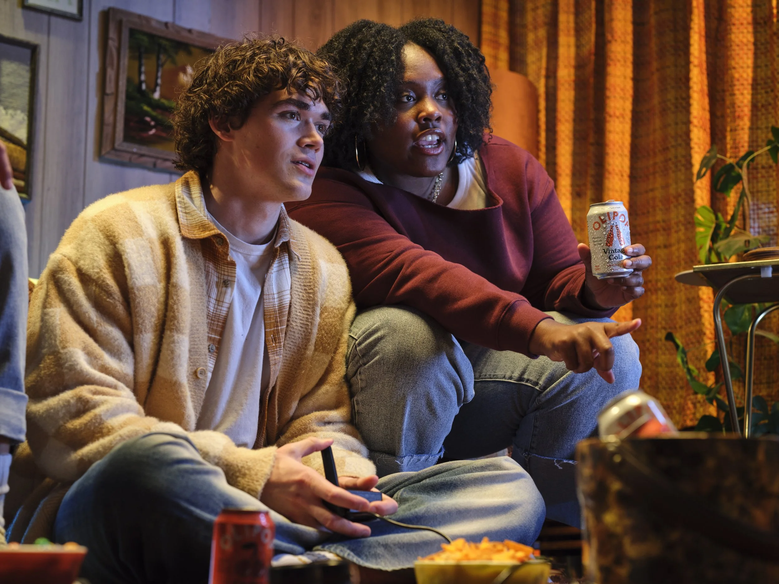 Two people sitting on a couch, engaged in an activity. The woman on the right is holding a can of soda and pointing at something, while the man on the left is holding a game controller or remote. The scene has a cozy, retro vibe with warm colors and framed pictures on the wall.